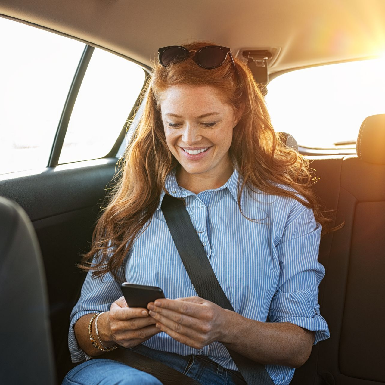 Une femme assise à l'arrière d'une voiture sourit en utilisant son téléphone, portant sa ceinture de sécurité.