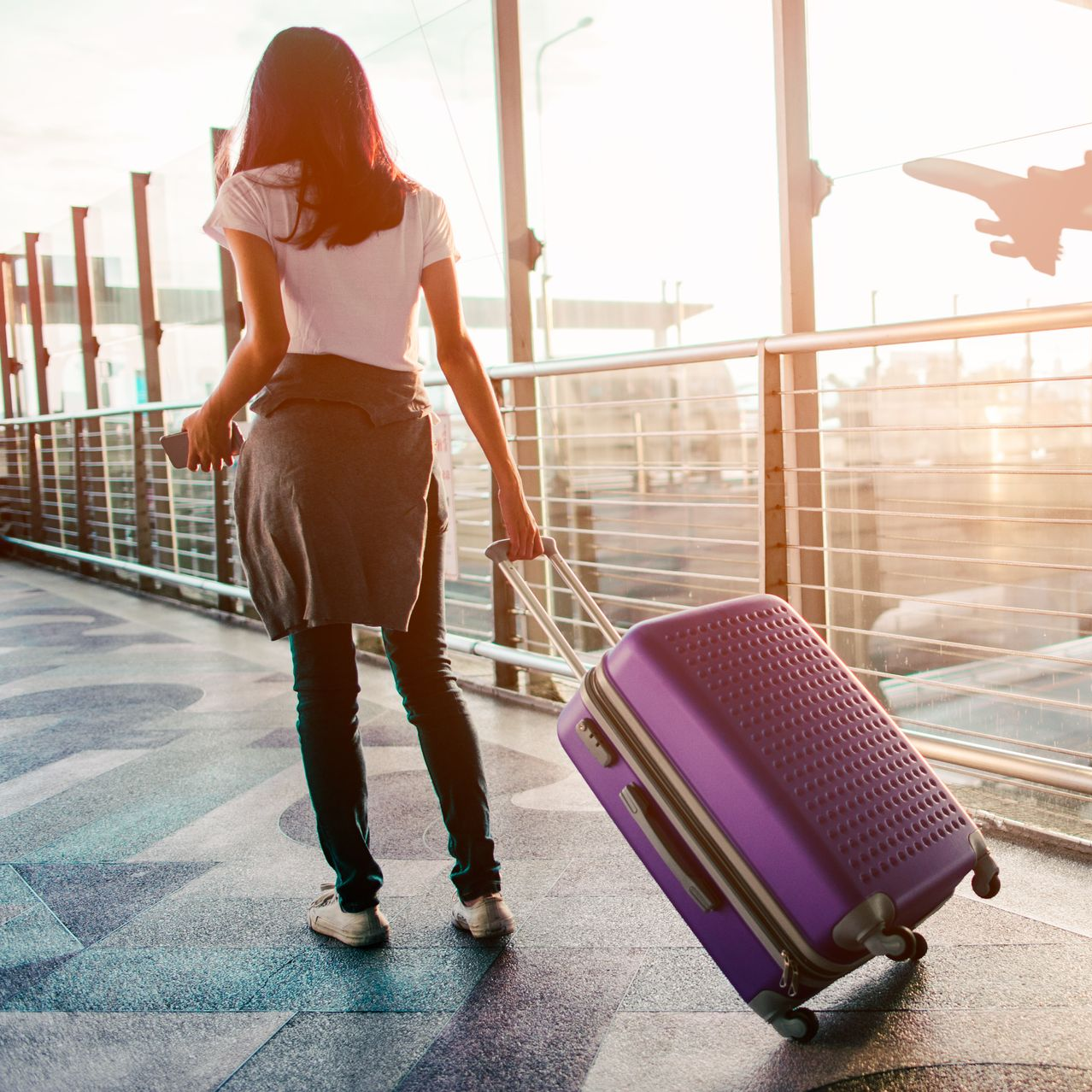 Une femme traverse un aéroport en tirant une valise violette, un avion étant visible en arrière-plan.