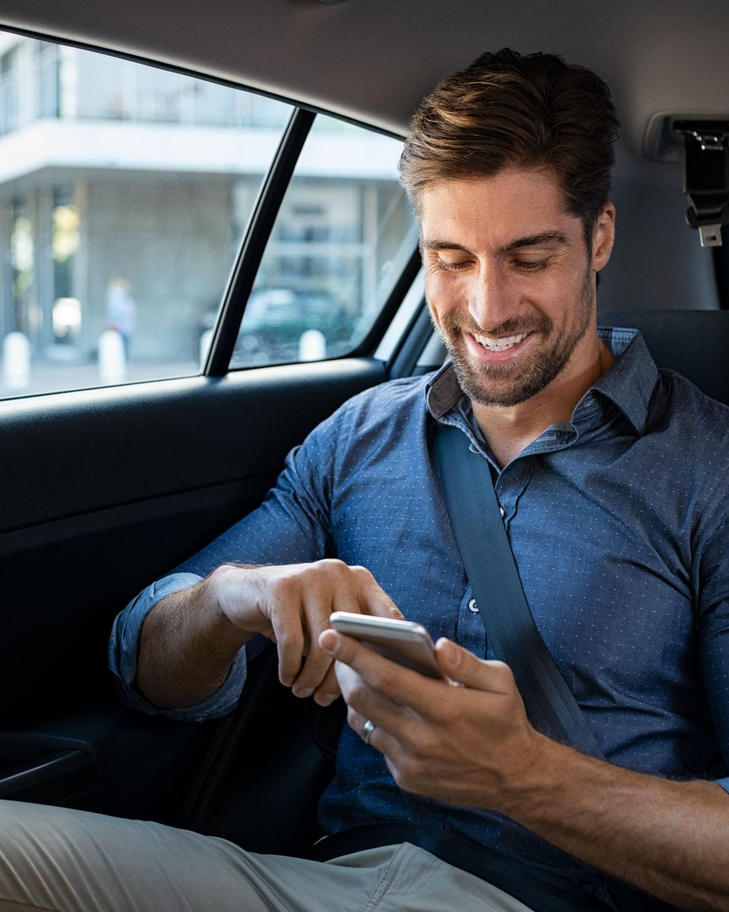 Un homme en chemise bleue, souriant, utilise son téléphone en étant assis dans une voiture.