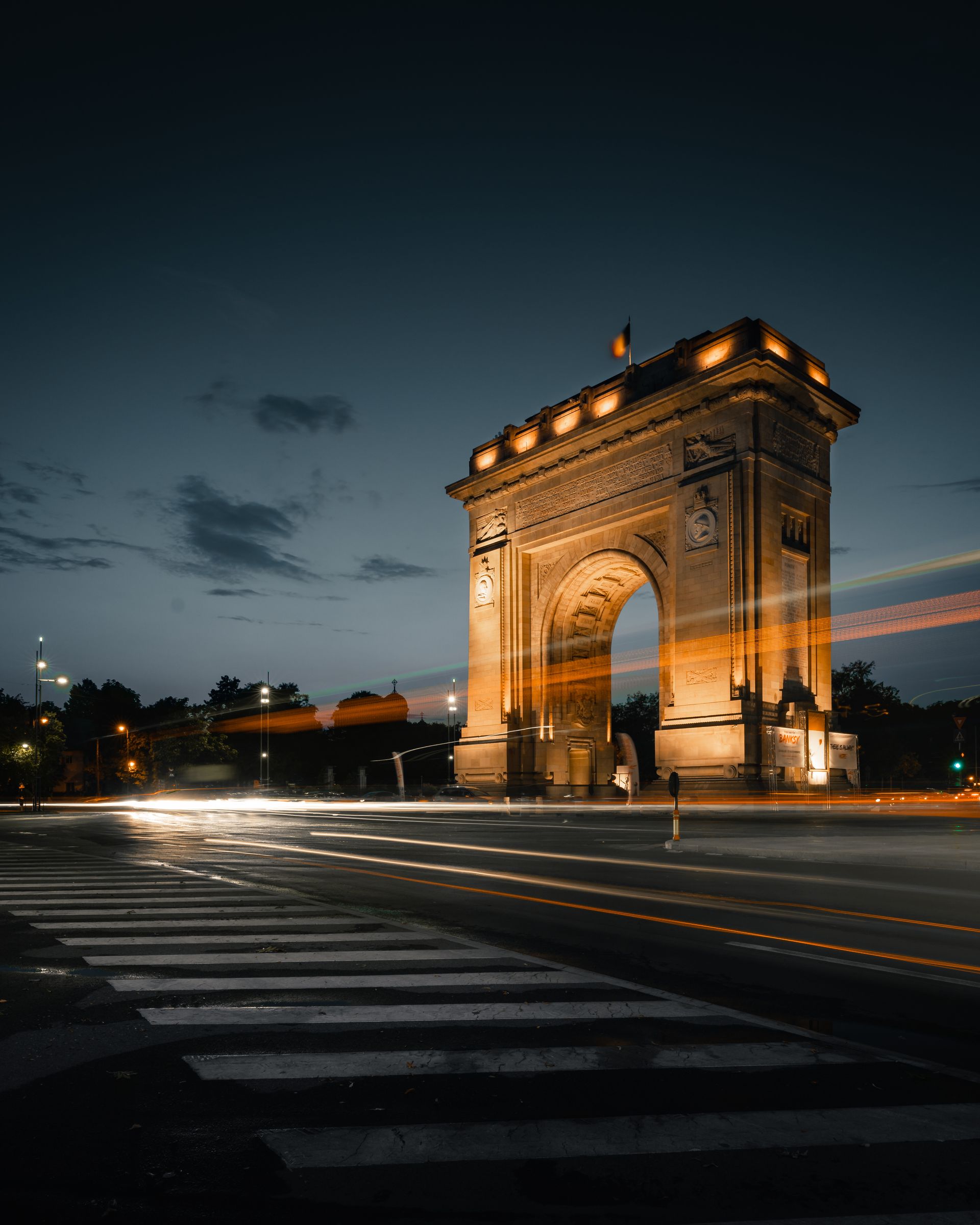 L'Arc de Triomphe illuminé la nuit, à Paris. Lumières orangées sur la structure et faisceaux de phares sur la chaussée mouillée.