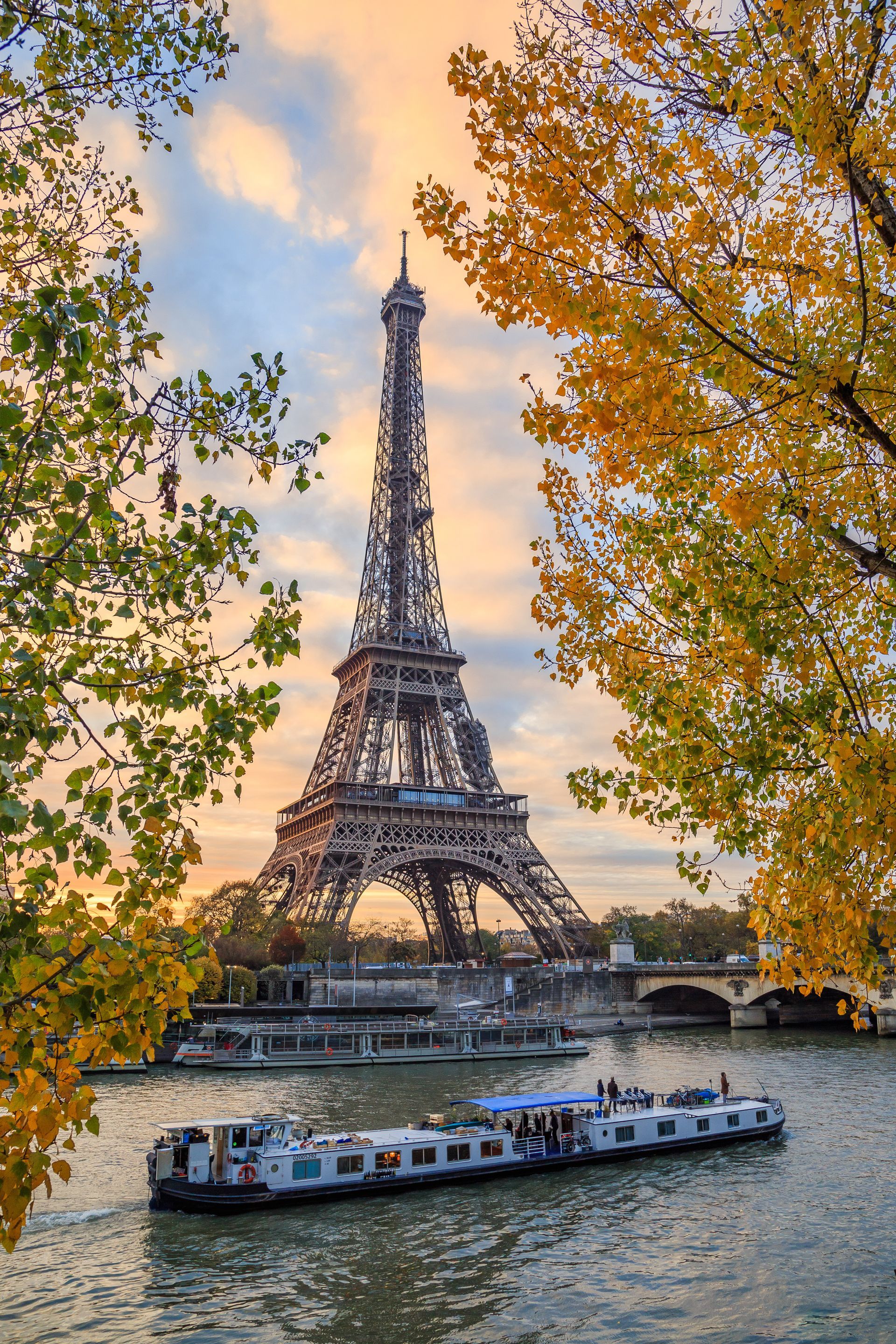 La tour Eiffel à Paris, encadrée par les arbres d'automne surplombant la Seine, avec des bateaux qui passent.