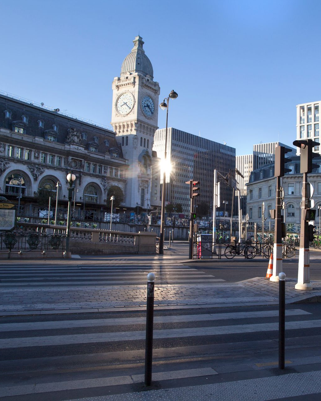 Vue de rue d'une tour d'horloge et d'immeubles. La lumière du soleil traverse les bâtiments, créant des reflets. Passage piéton au premier plan.