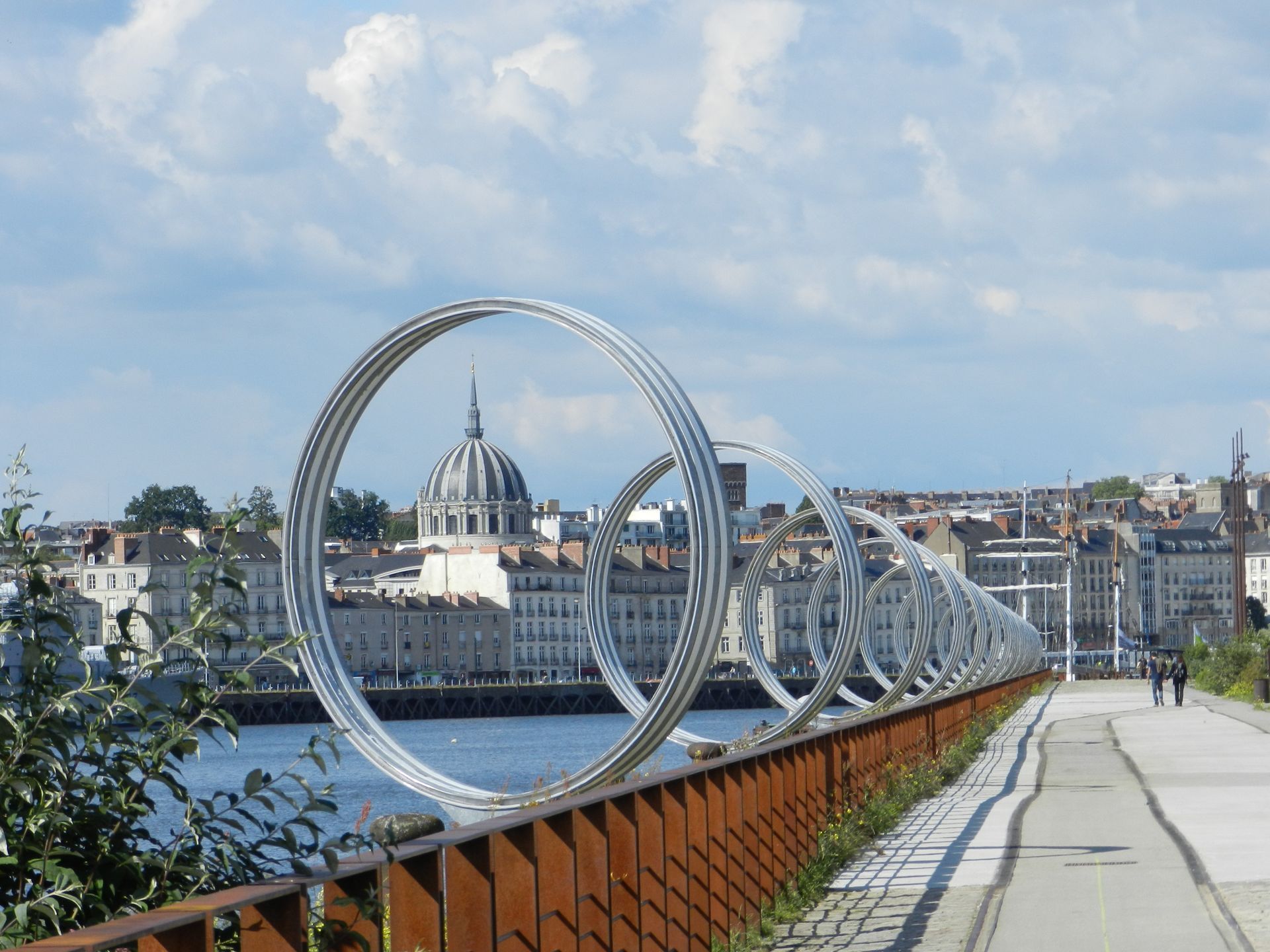Anneaux métalliques sculpturaux le long d'une promenade en bord de mer, paysage urbain et bâtiment à dôme en arrière-plan.