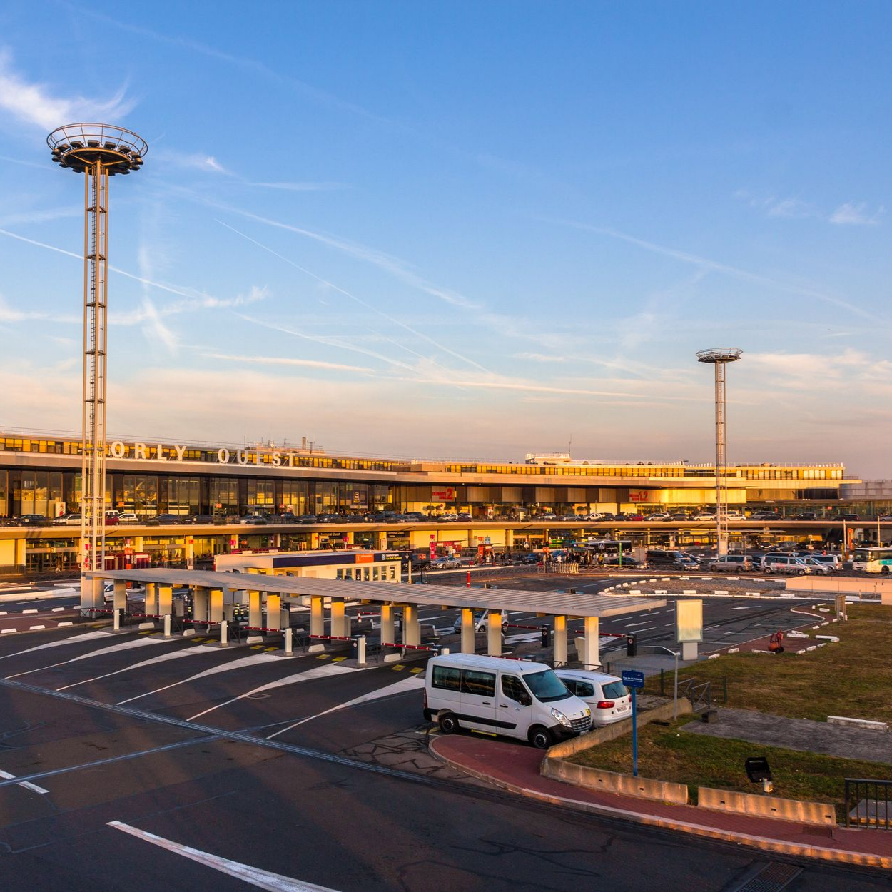 Extérieur d'un terminal d'aéroport avec une tour de contrôle, des véhicules stationnés à l'extérieur et un ciel bleu dégagé.