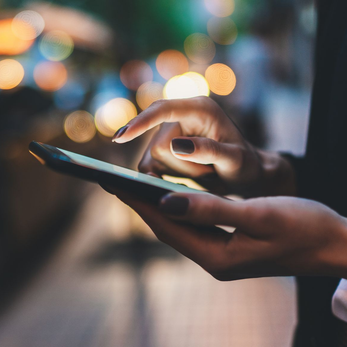 Des mains utilisant un smartphone, illuminées par la lueur de l'écran, avec des lumières floues (effet bokeh) en arrière-plan.