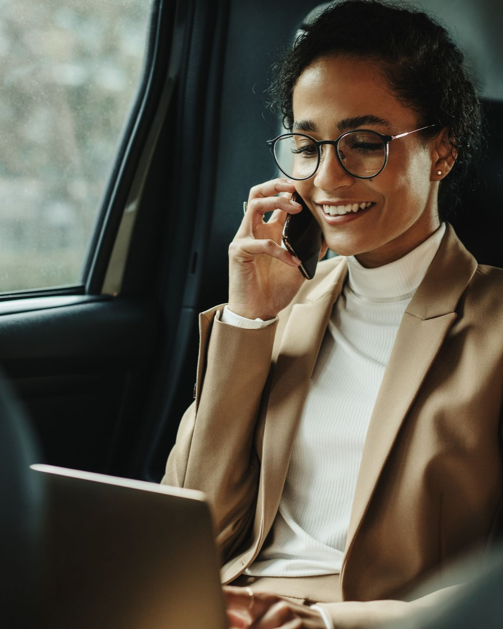 Une femme dans une voiture, souriante tout en étant au téléphone et en regardant un ordinateur portable, portant des lunettes.