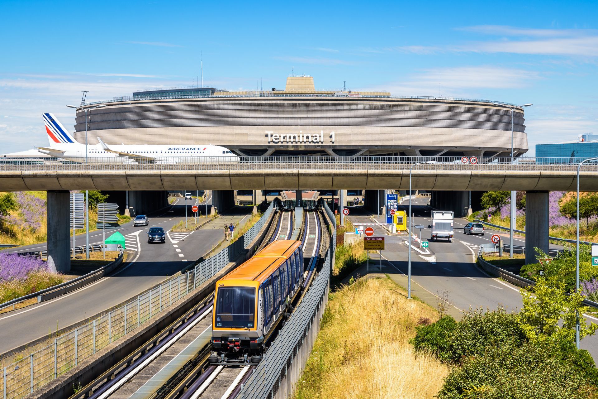 Train circulant sur des voies ferrées avec un terminal d'aéroport, un avion et une route au-dessus. Ciel bleu et ensoleillé.