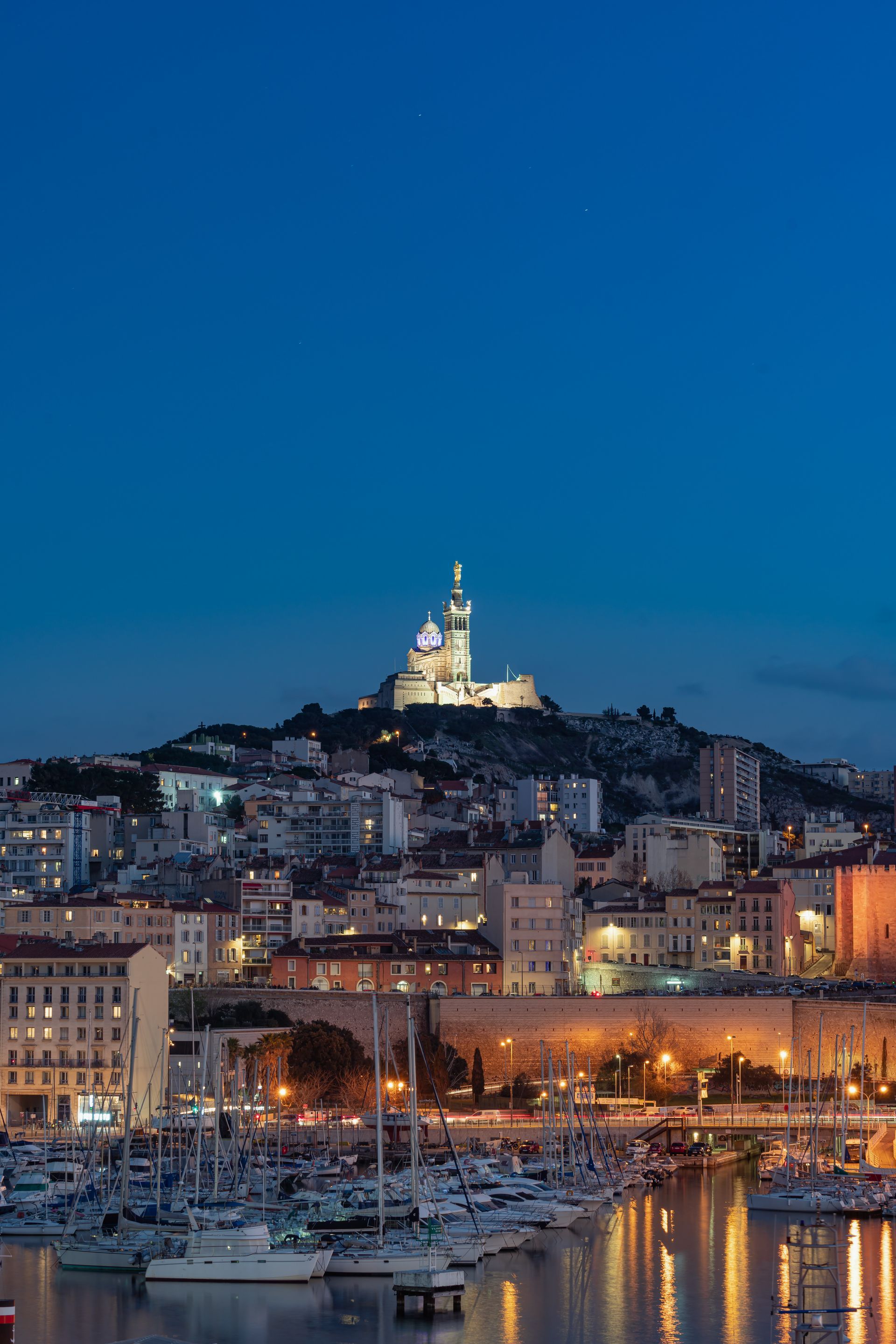 Vue nocturne de Marseille, en France, avec des bateaux dans le port et la basilique Notre-Dame de la Garde illuminée sur une colline.