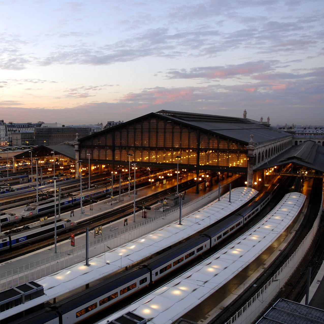 Gare au crépuscule, intérieur éclairé, plusieurs voies ferrées avec des trains. Bâtiments en arrière-plan, ciel nuageux.