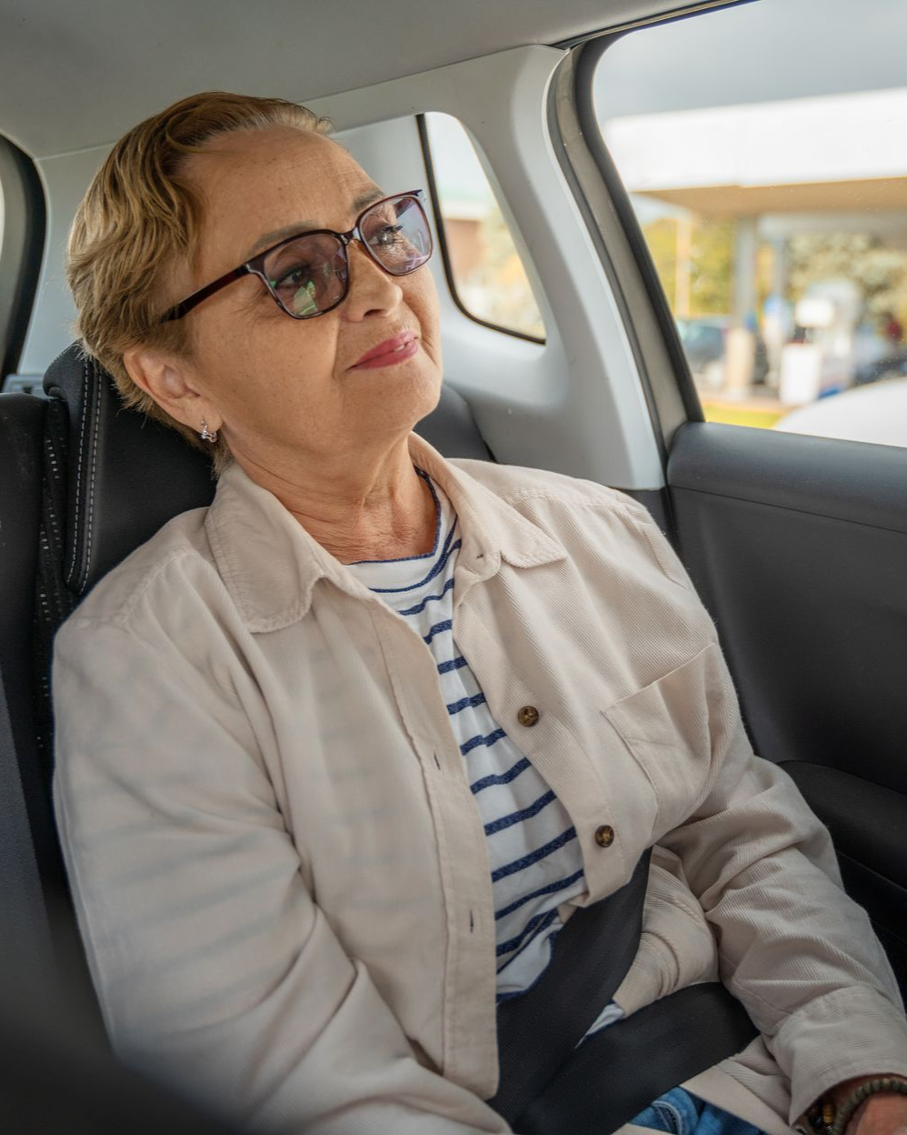 Femme portant des lunettes, une chemise à rayures et une veste légère, assise dans une voiture, regardant par la fenêtre.