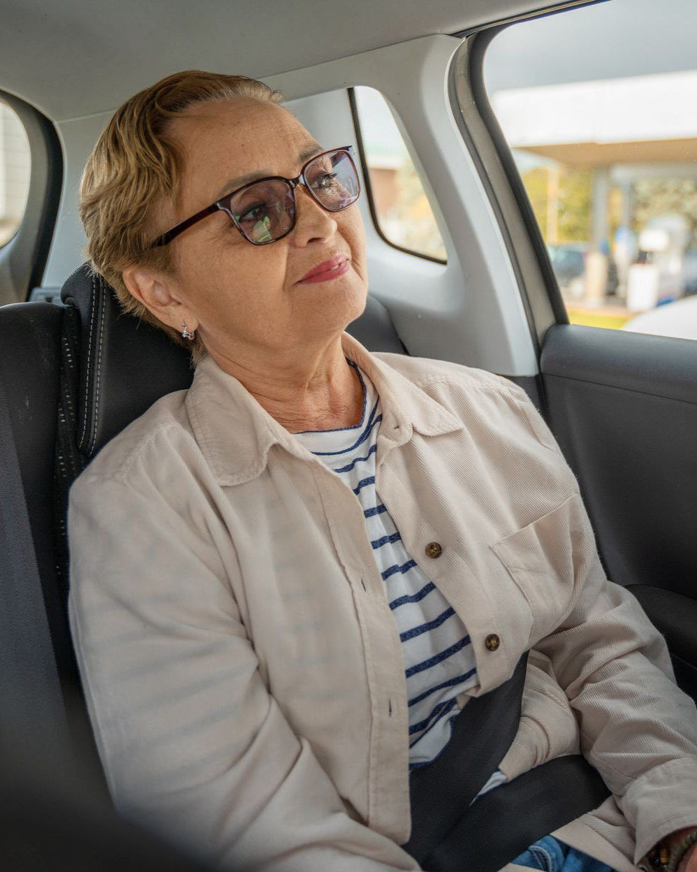 Une femme portant des lunettes et une veste beige, assise sur la banquette arrière d'une voiture, regarde vers le haut et sourit.
