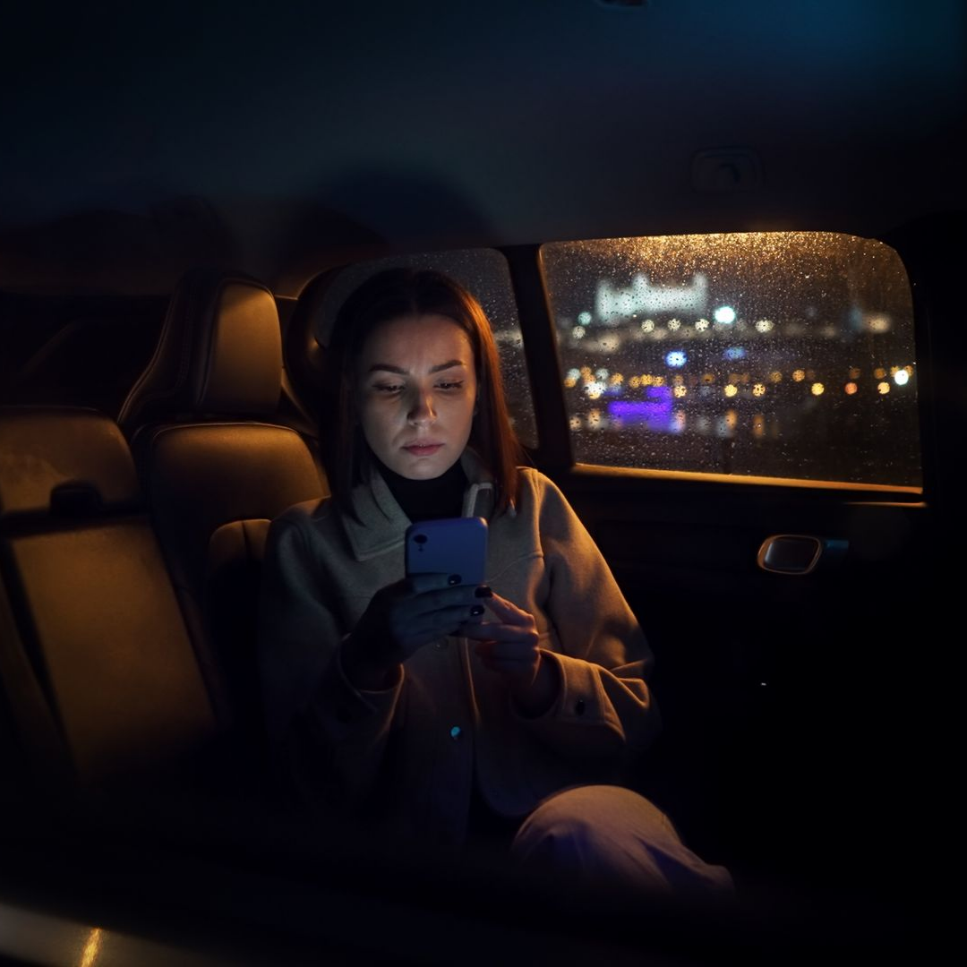 Une femme en voiture utilise son téléphone la nuit, les lumières de la ville sont visibles à travers la vitre.