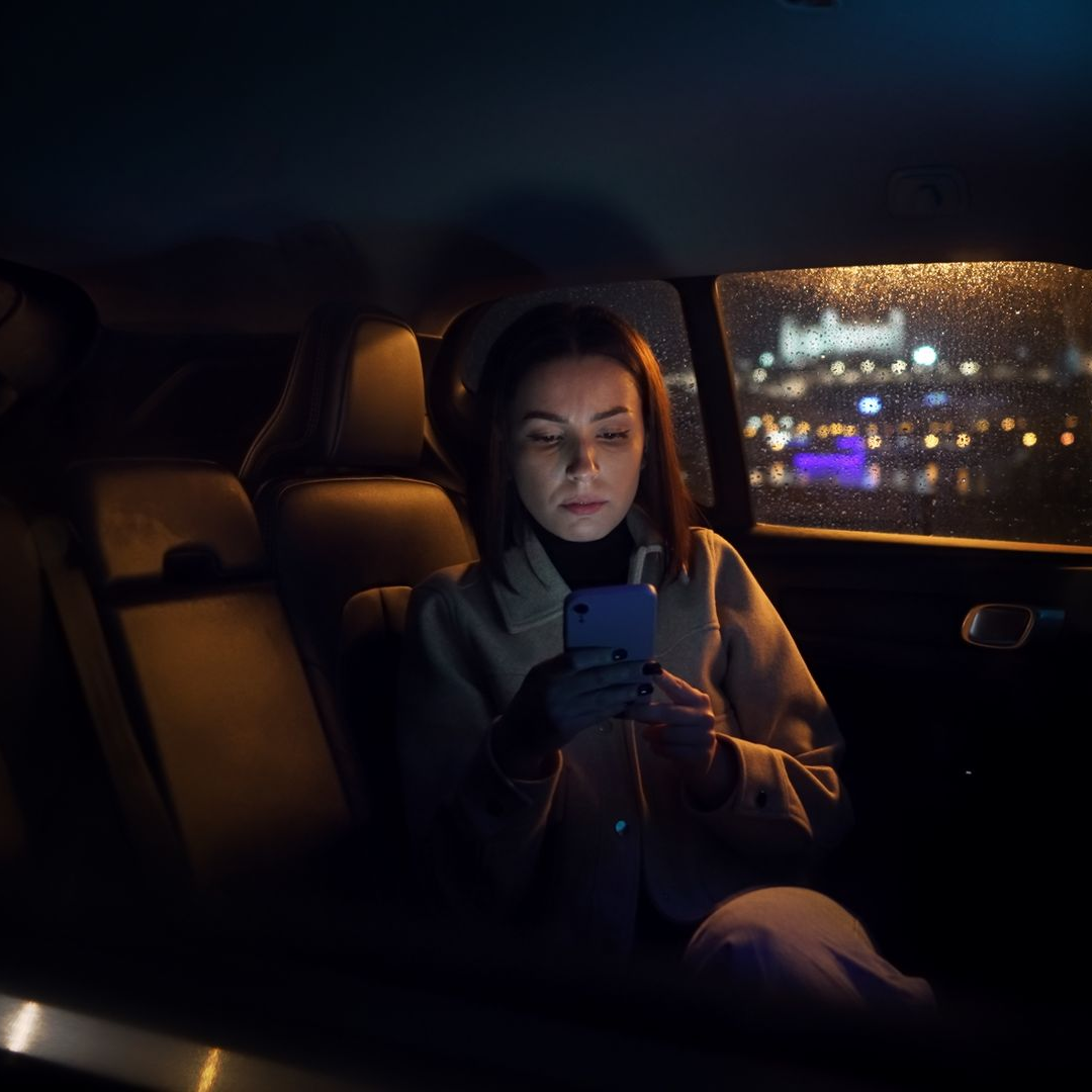 Une femme utilise son téléphone sur la banquette arrière d'une voiture, de nuit, les yeux rivés sur l'écran. On aperçoit les lumières de la ville au loin.