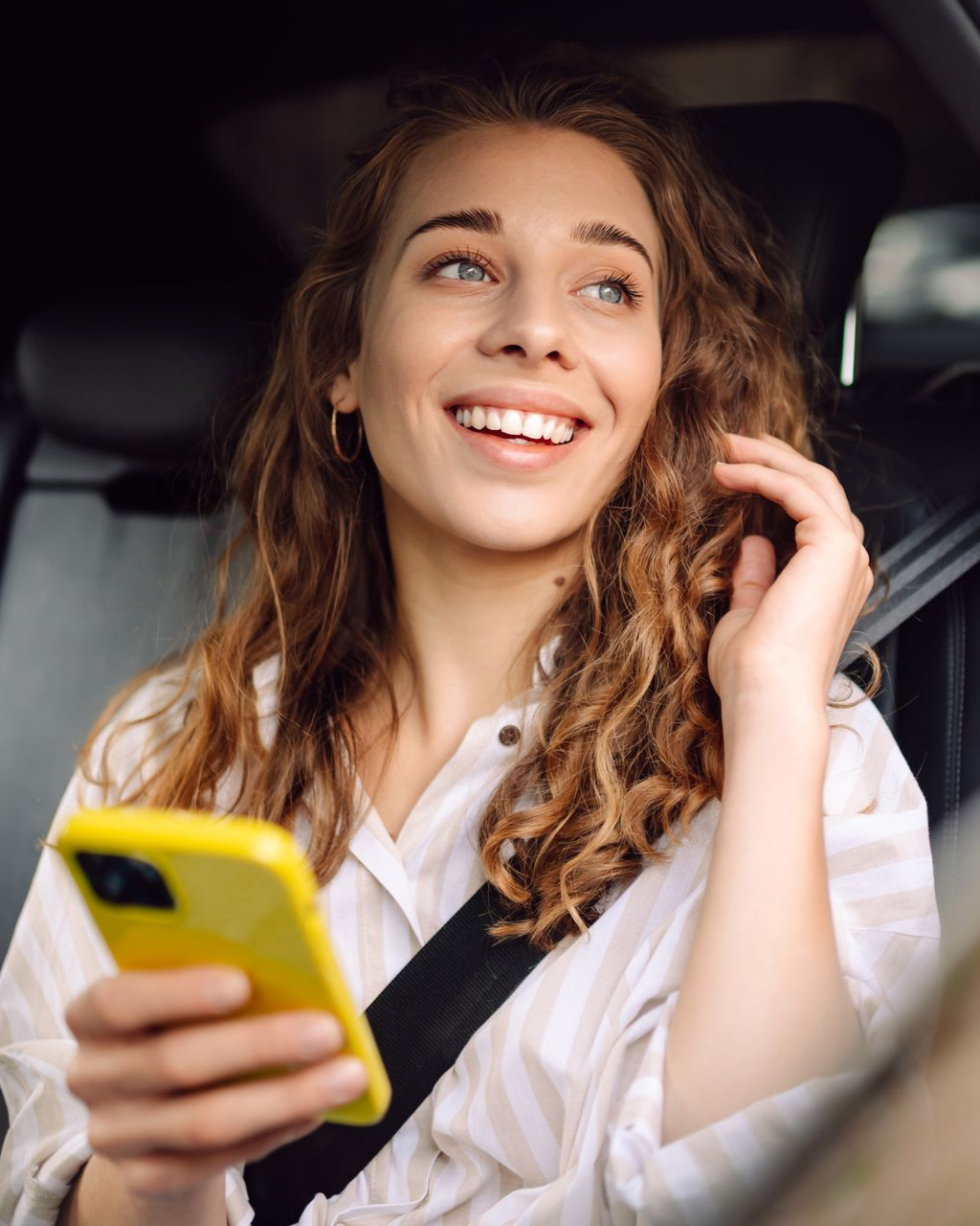 Femme souriante, tenant un téléphone jaune, portant sa ceinture de sécurité à l'intérieur d'une voiture.