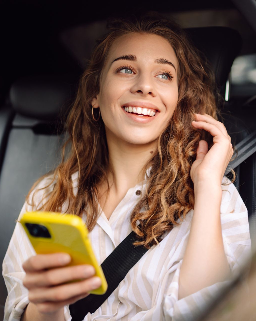 Femme souriante, tenant un téléphone jaune dans une voiture, regardant vers le haut et la droite. Elle porte une chemise à rayures blanches et sa ceinture de sécurité est attachée.