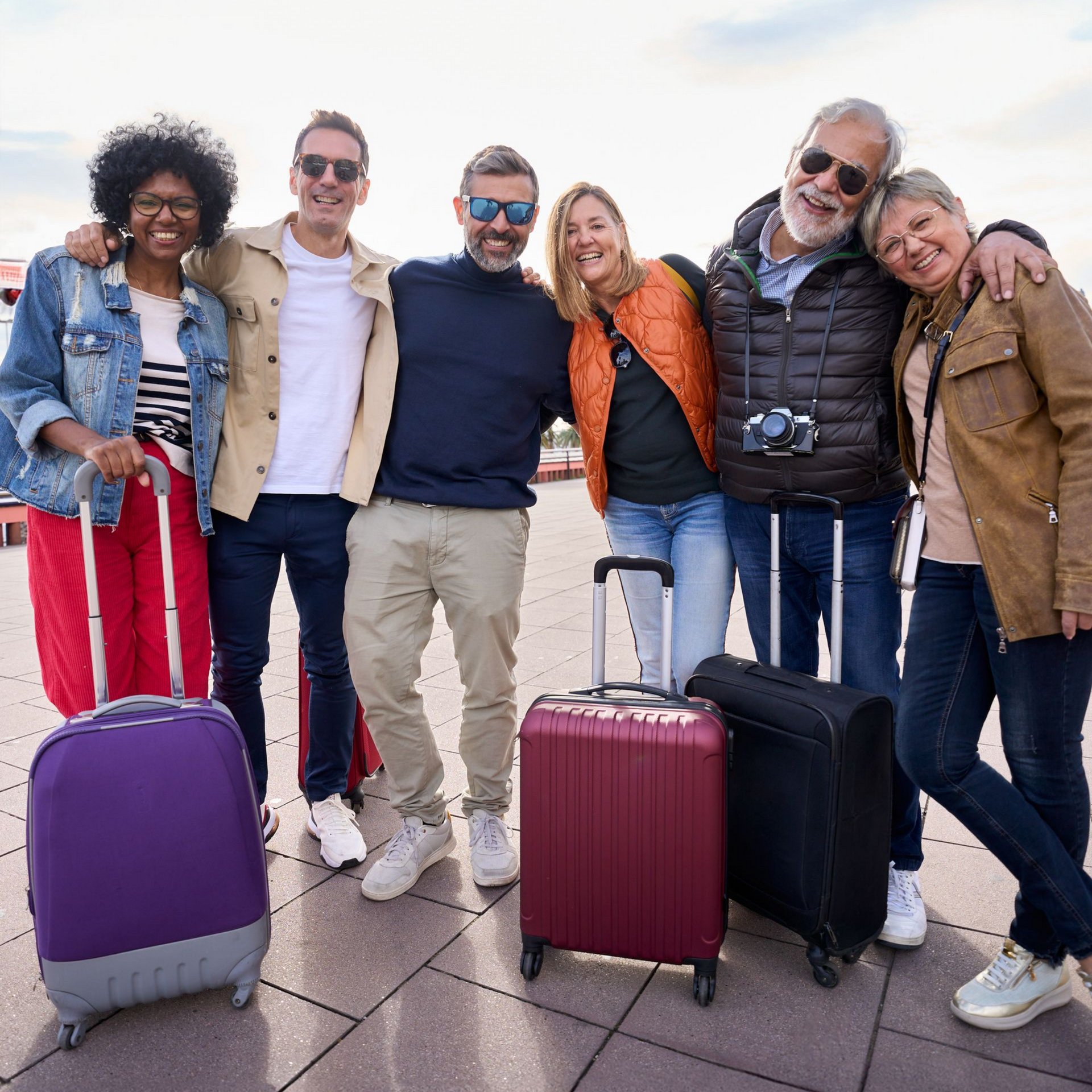 Un groupe de personnes souriantes avec leurs bagages, probablement en vacances, posant à l'extérieur par une journée ensoleillée.