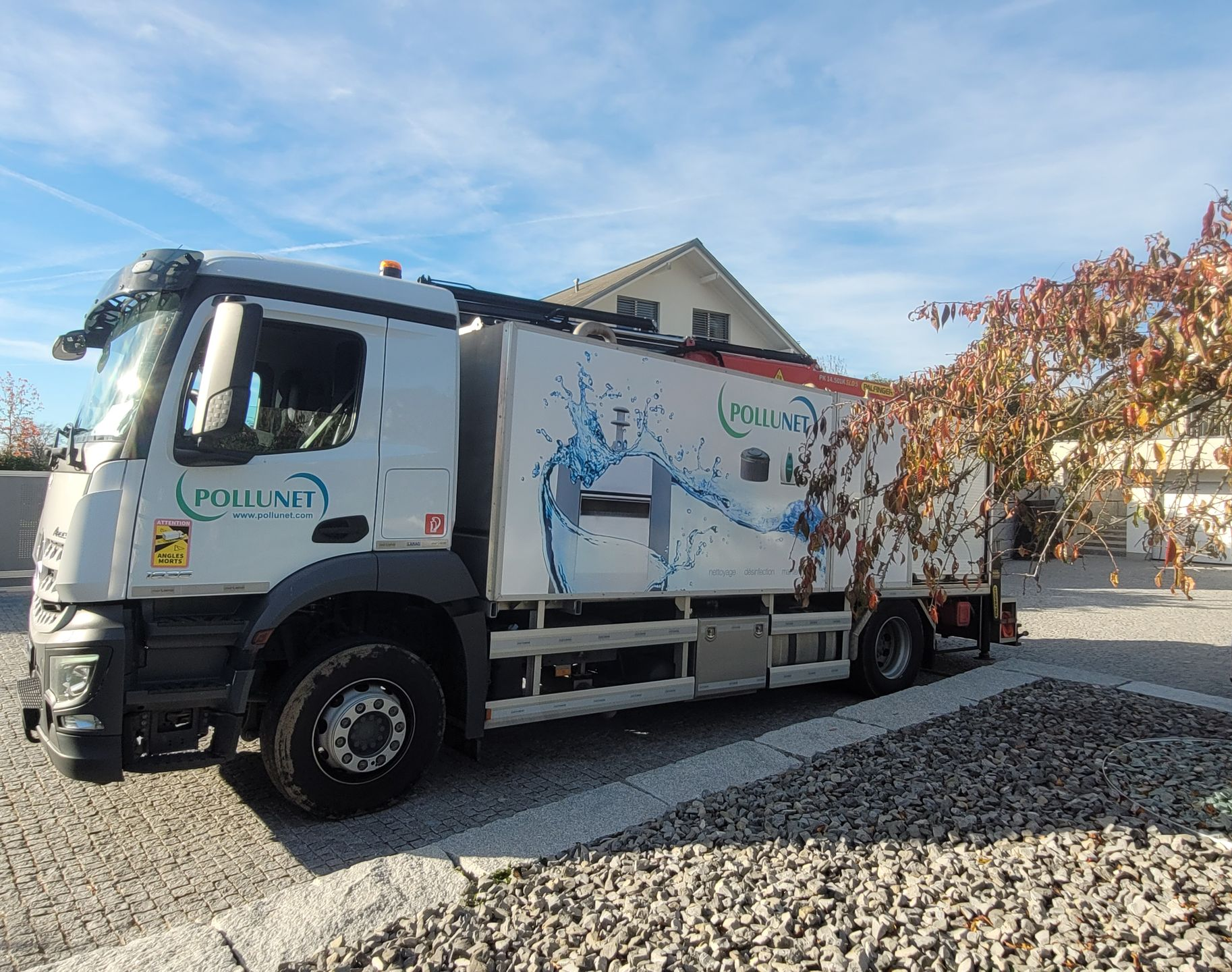 Un camion avec une grue vide un bac de recyclage devant un bâtiment ; un ouvrier en gilet orange au travail.