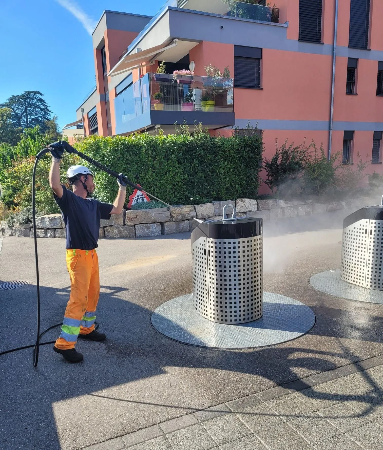Un homme en orange nettoie une poubelle avec un nettoyeur haute pression. Bâtiments et arbustes en arrière-plan.