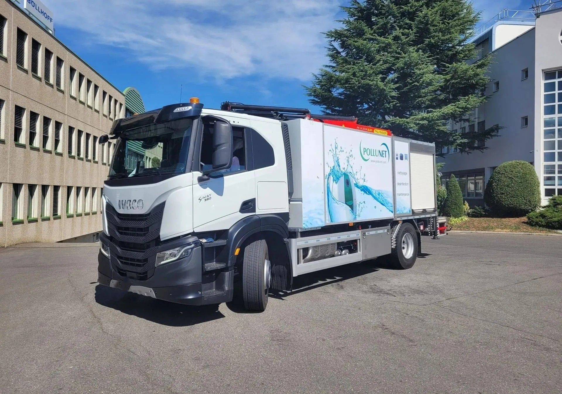 Camion Iveco blanc garé devant un bâtiment avec un motif d'eau bleu et blanc sur le côté. Journée ensoleillée.