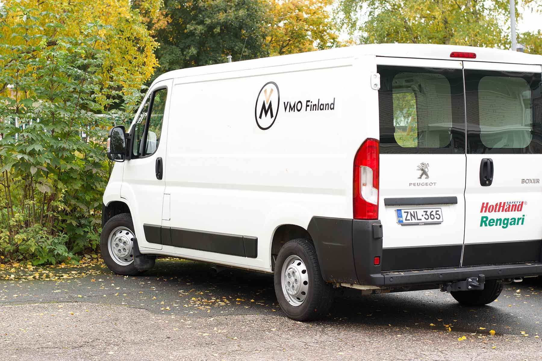 Valkoinen pakettiauto pysäköitynä sorakentälle syksyisten puiden viereen, kyljessä värikkäät logot.