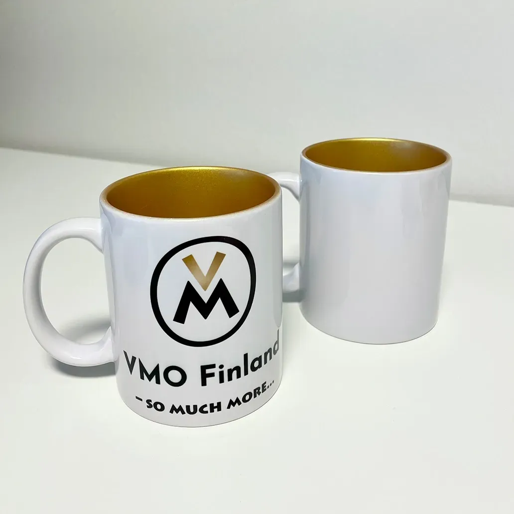 Kaksi valkoista mukia, joissa on kultainen sisus; toisessa on YMO Finland -logo valkoisella taustalla.