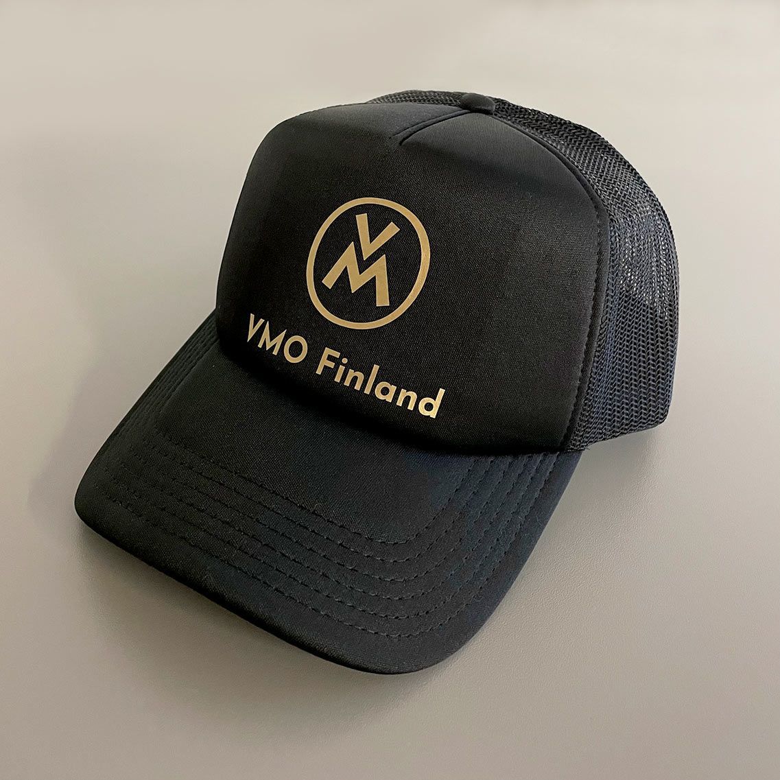 Musta verkkokankainen lippalakki, jossa edessä kultainen ”VMO Finland” -logo, kulmassa vaalealla pinnalla