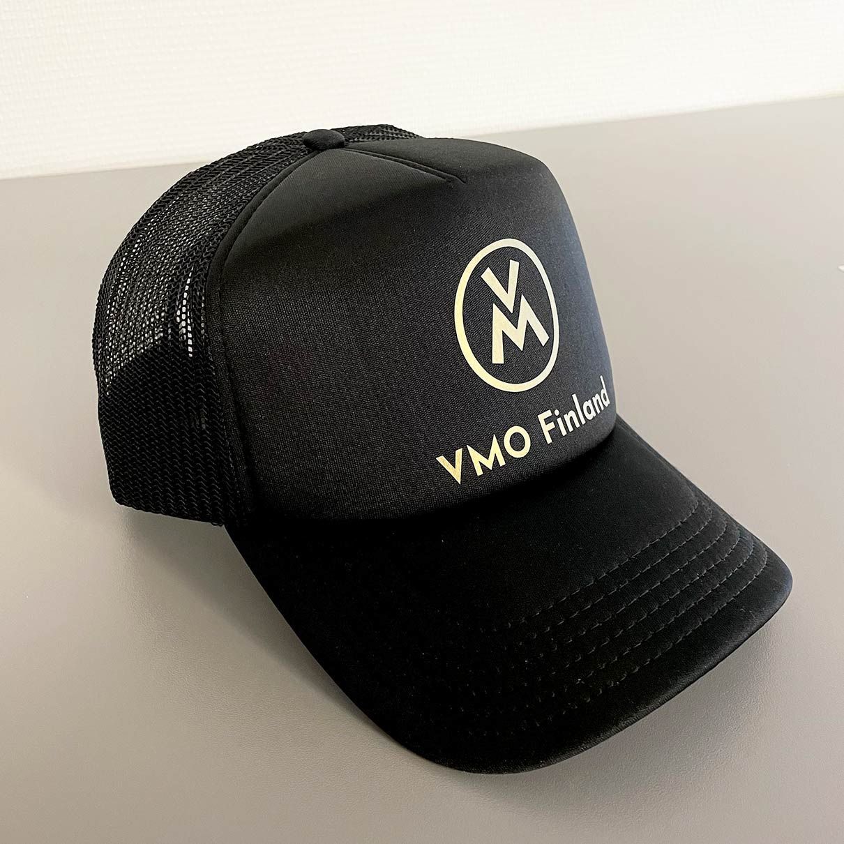 Musta verkkokankainen rekkakuskin lippis, jossa valkoinen pyöreä logo ja teksti ”VMO” harmaalla pinnalla