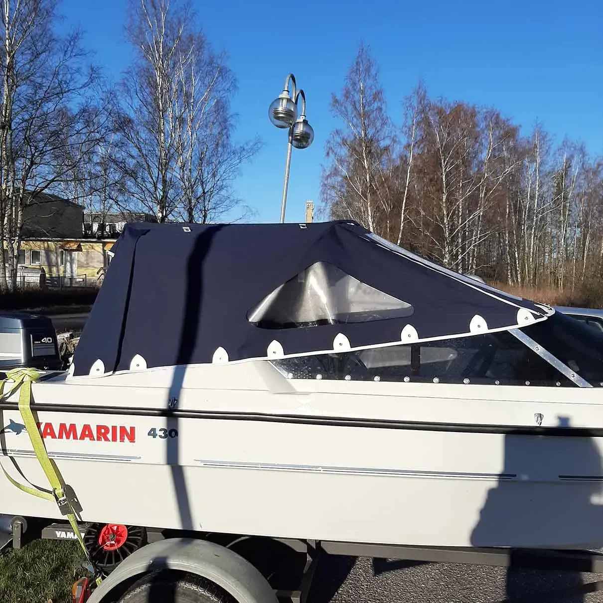 Pieni valkoinen vene, peitetty tummalla pressulla, pysäköitynä ulkona sinisen taivaan alle paljaiden puiden lähellä.