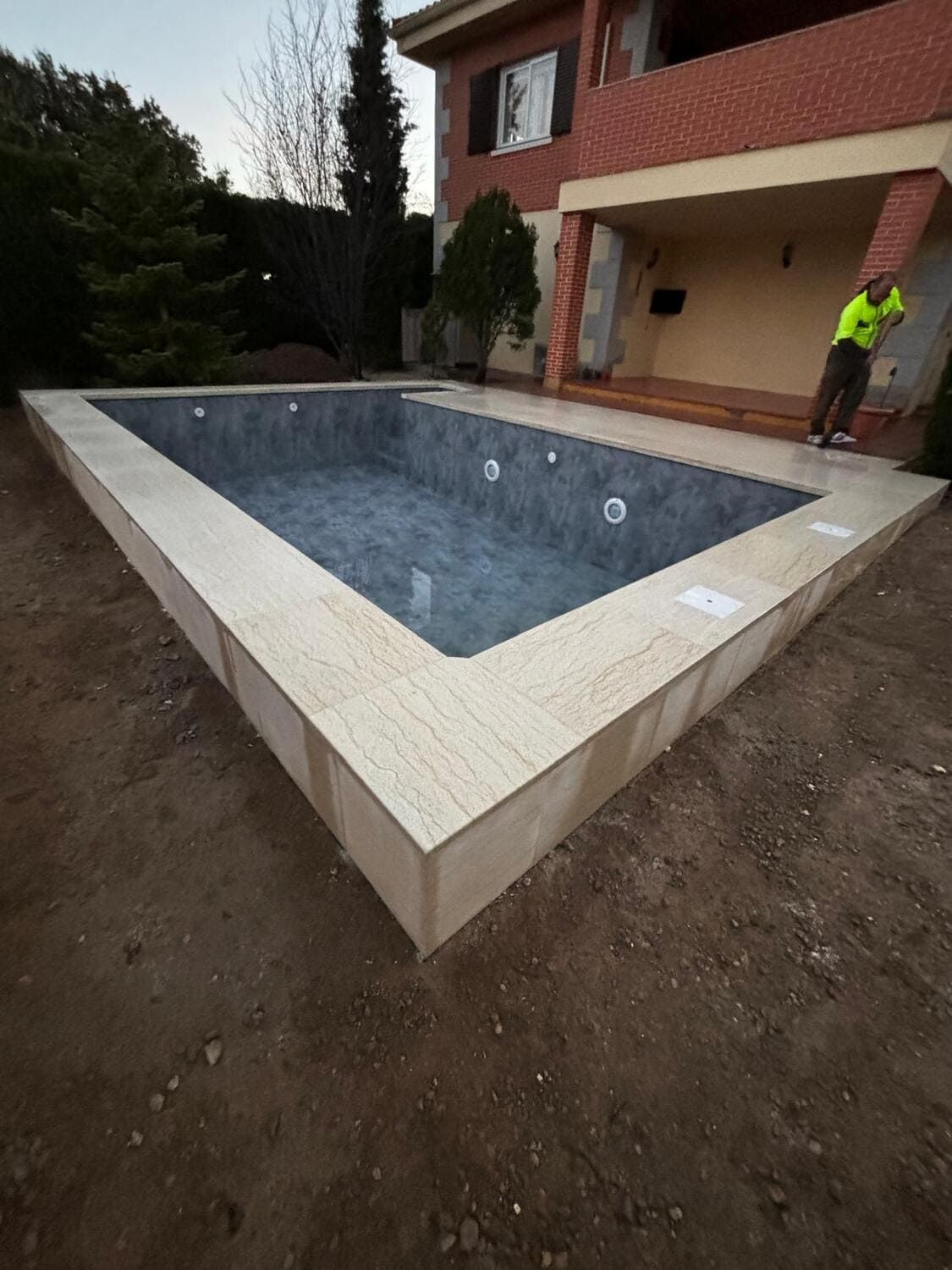 Piscina rectangular vacía con borde de piedra frente a una casa de ladrillo; una persona está de pie en el patio.