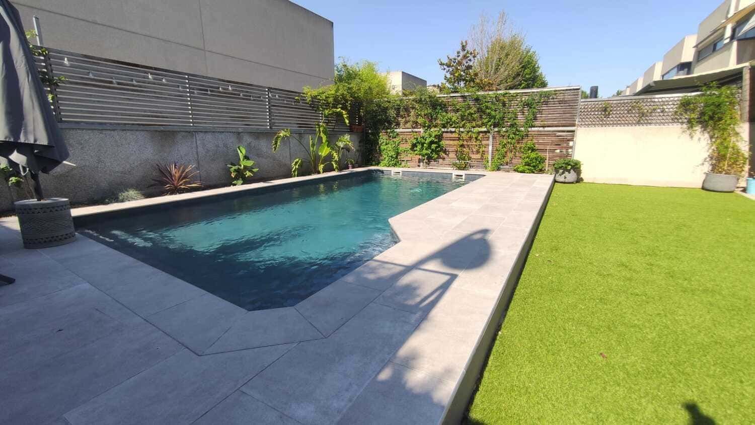 Piscina en el patio trasero con césped verde, patio gris y vegetación circundante.