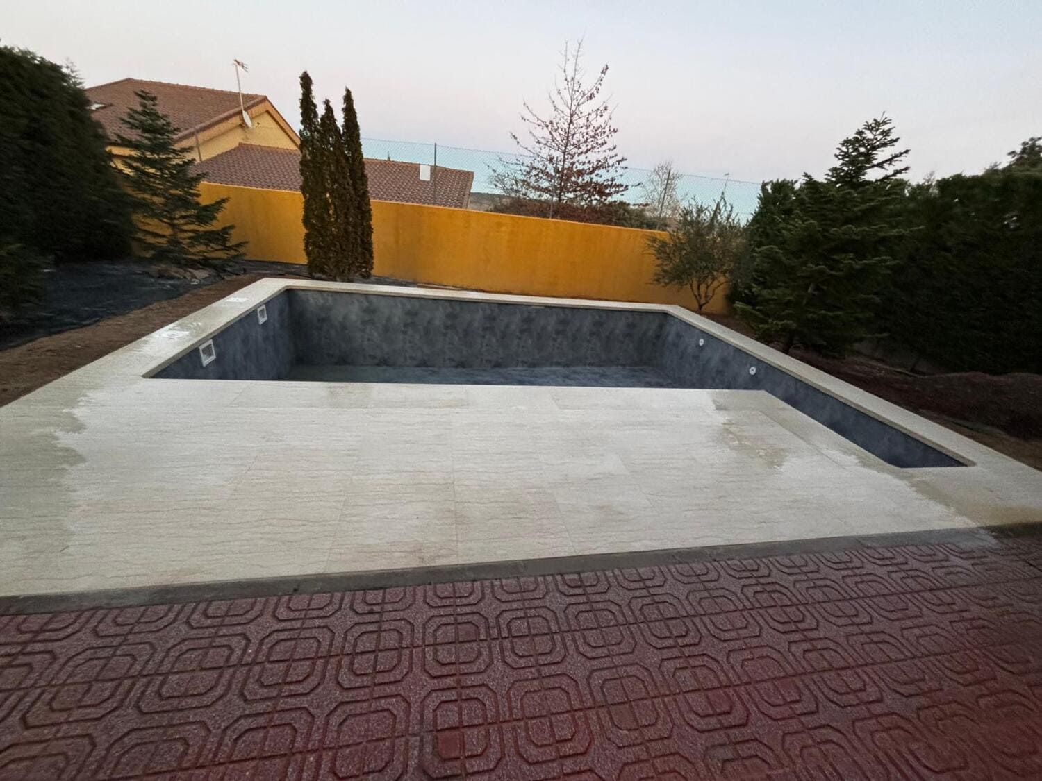 Piscina rectangular vacía con marco de hormigón, bordeada por un patio estampado en rojo.