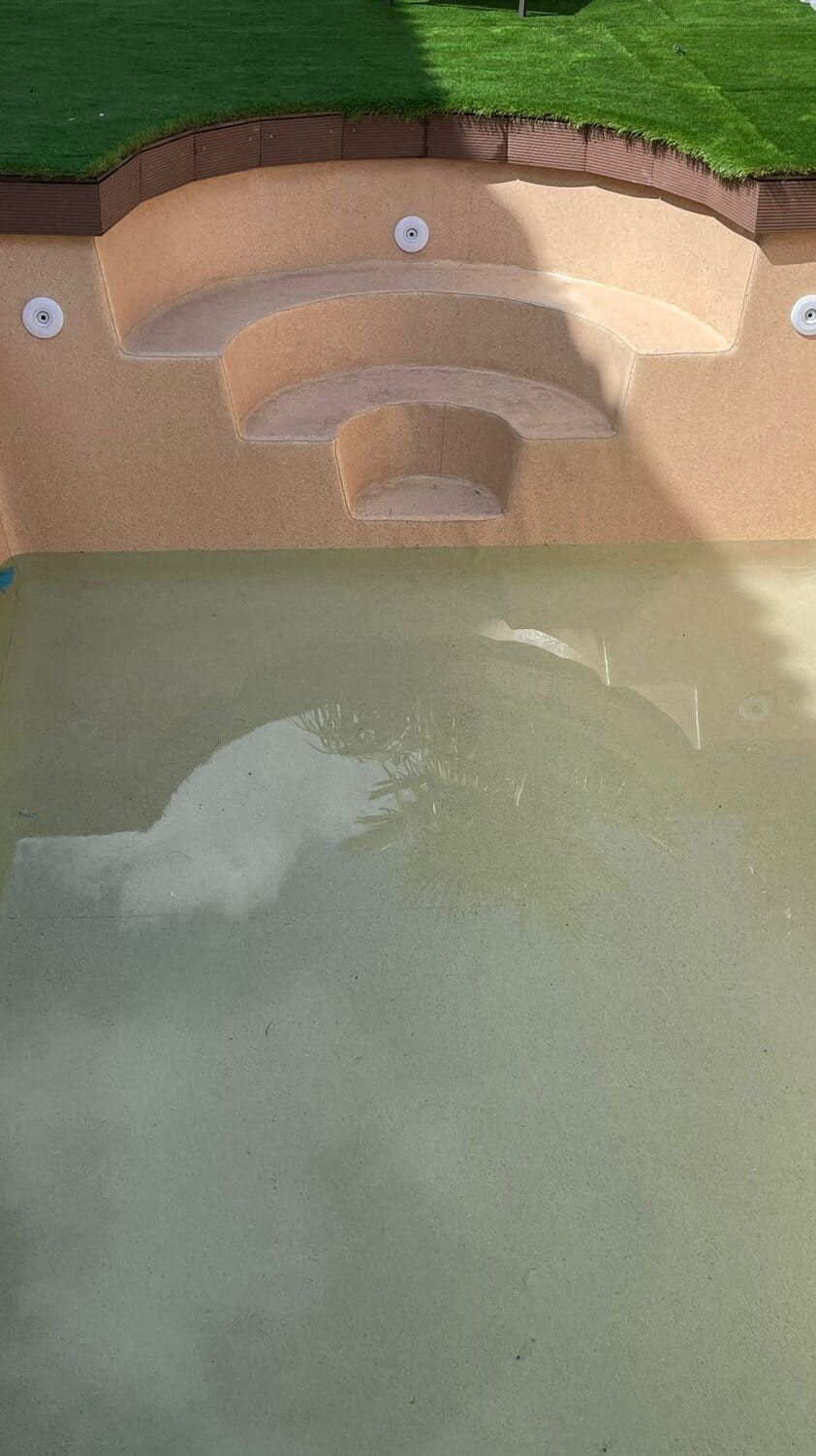 Piscina con escalones de color marrón claro y agua turbia, bordeada de césped verde y borde marrón.