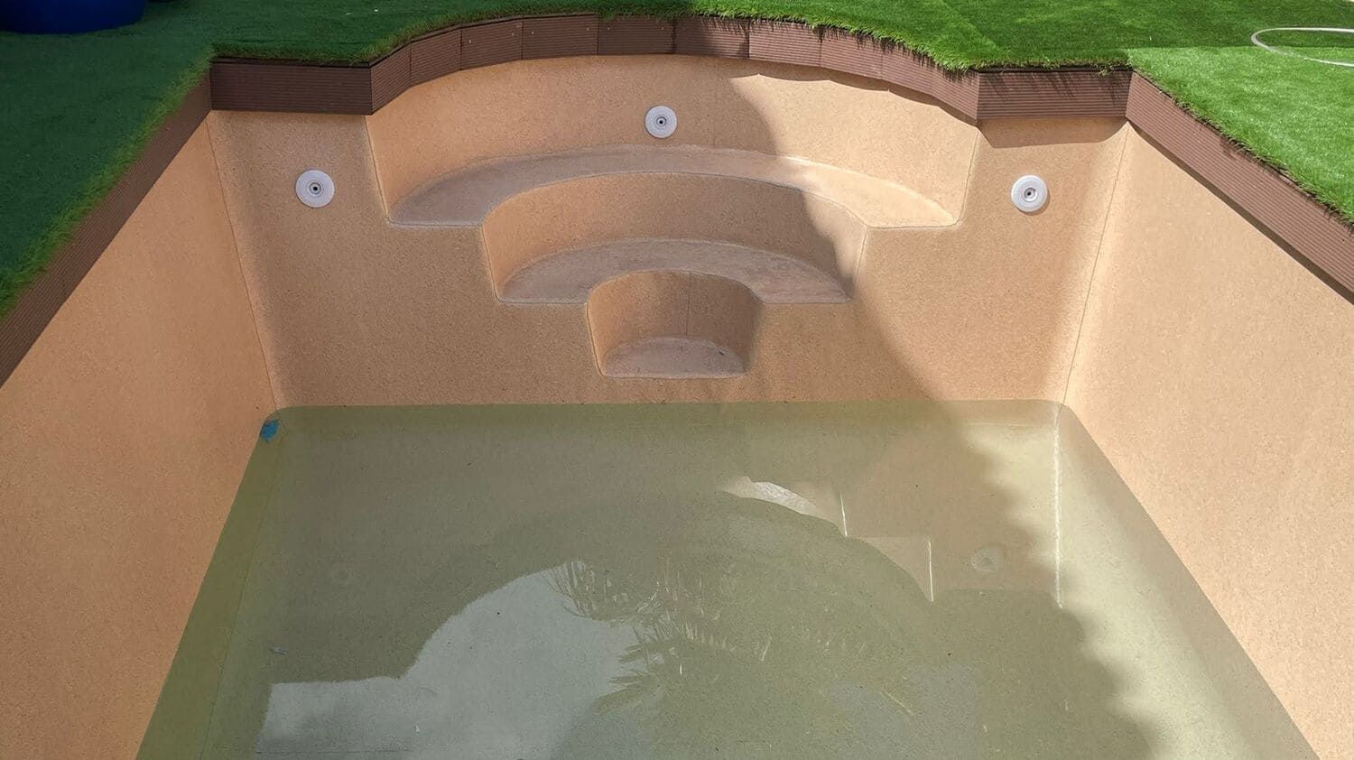 Piscina parcialmente llena con escalones. Paredes color canela y césped artificial alrededor.