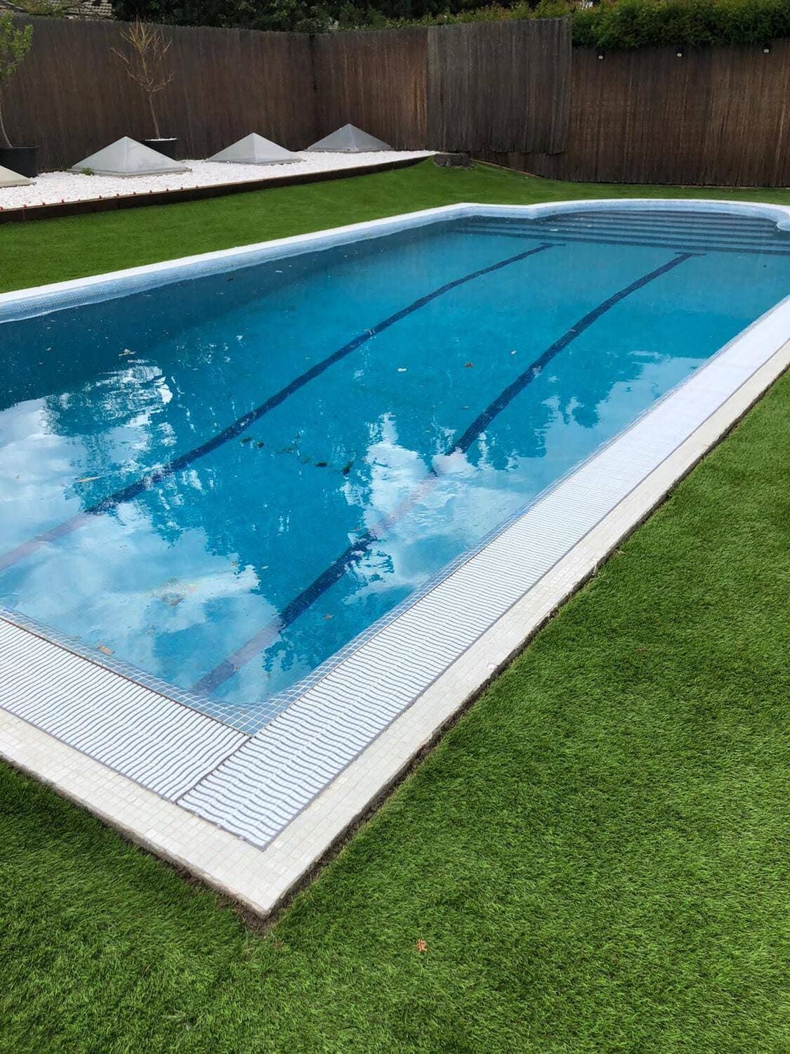 Piscina rectangular de agua azul y líneas de carril, rodeada de césped artificial verde.