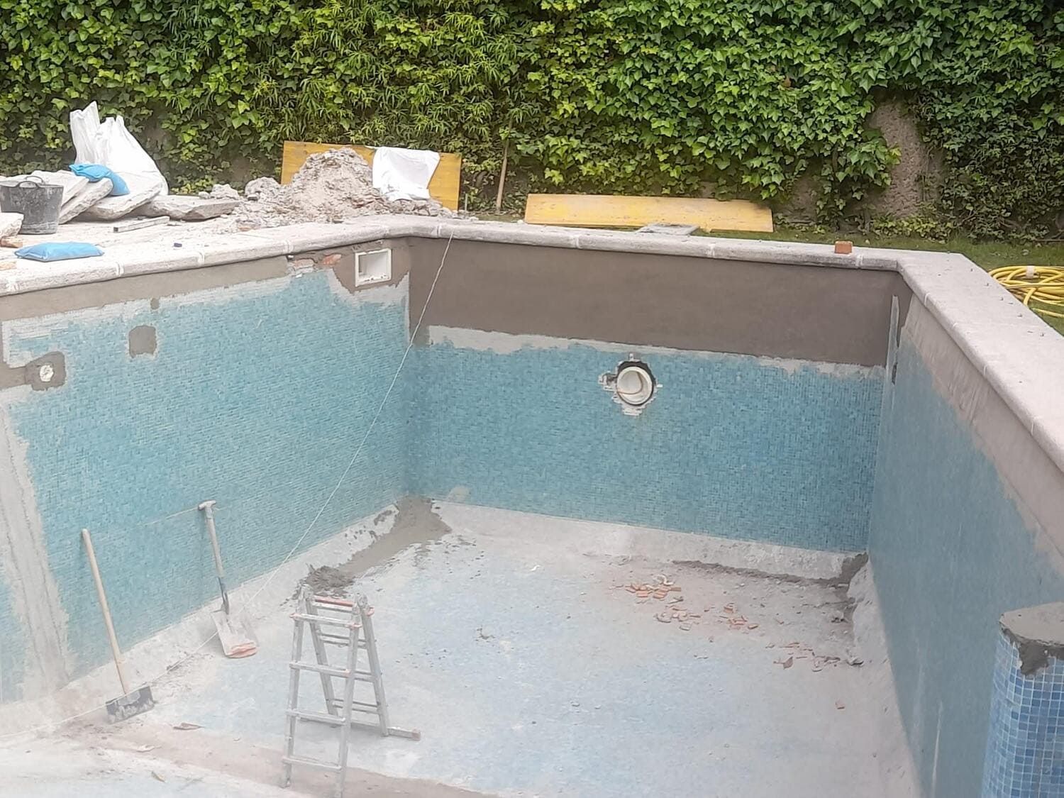 Piscina en construcción; paredes de azulejos turquesas, mortero gris, borde de hormigón, herramientas y escombros visibles.