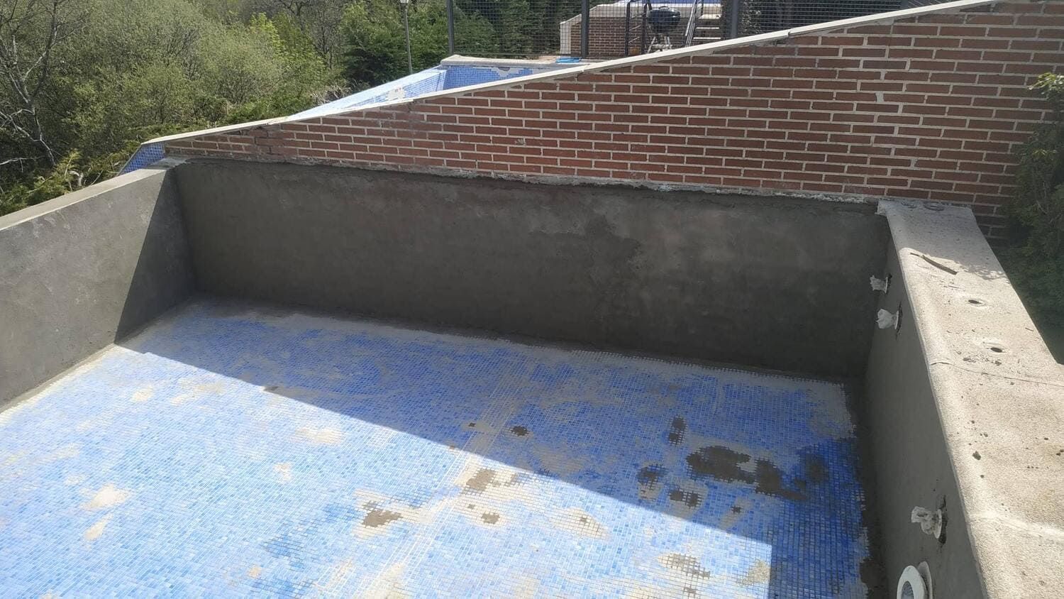 Piscina en construcción con suelo de baldosas azules, paredes de hormigón gris y pared de ladrillo al fondo.