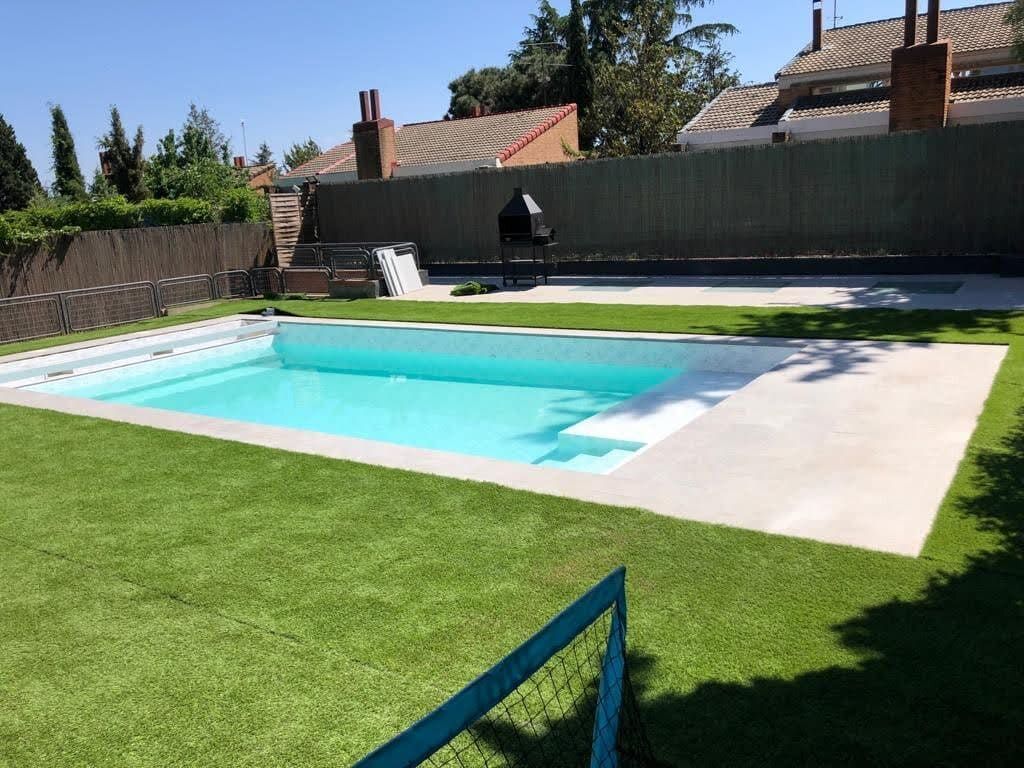 Una piscina rectangular rodeada de césped verde. Al fondo, una parrilla.