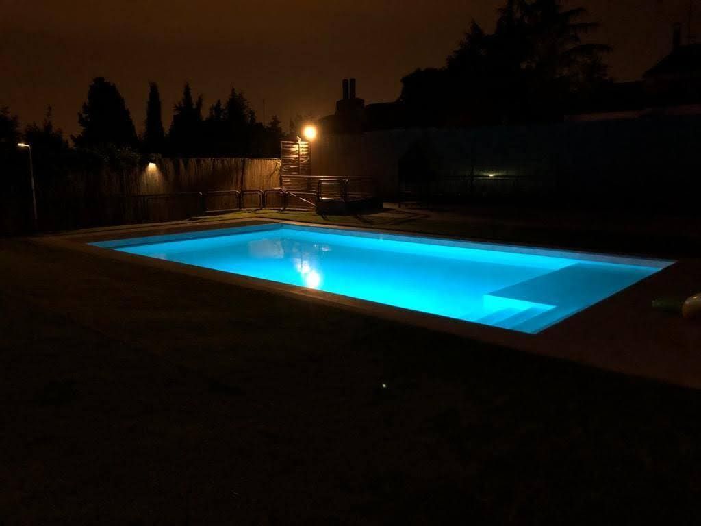 Vista nocturna de una piscina rectangular iluminada con luz azul. Entorno oscuro, árboles y edificios al fondo.