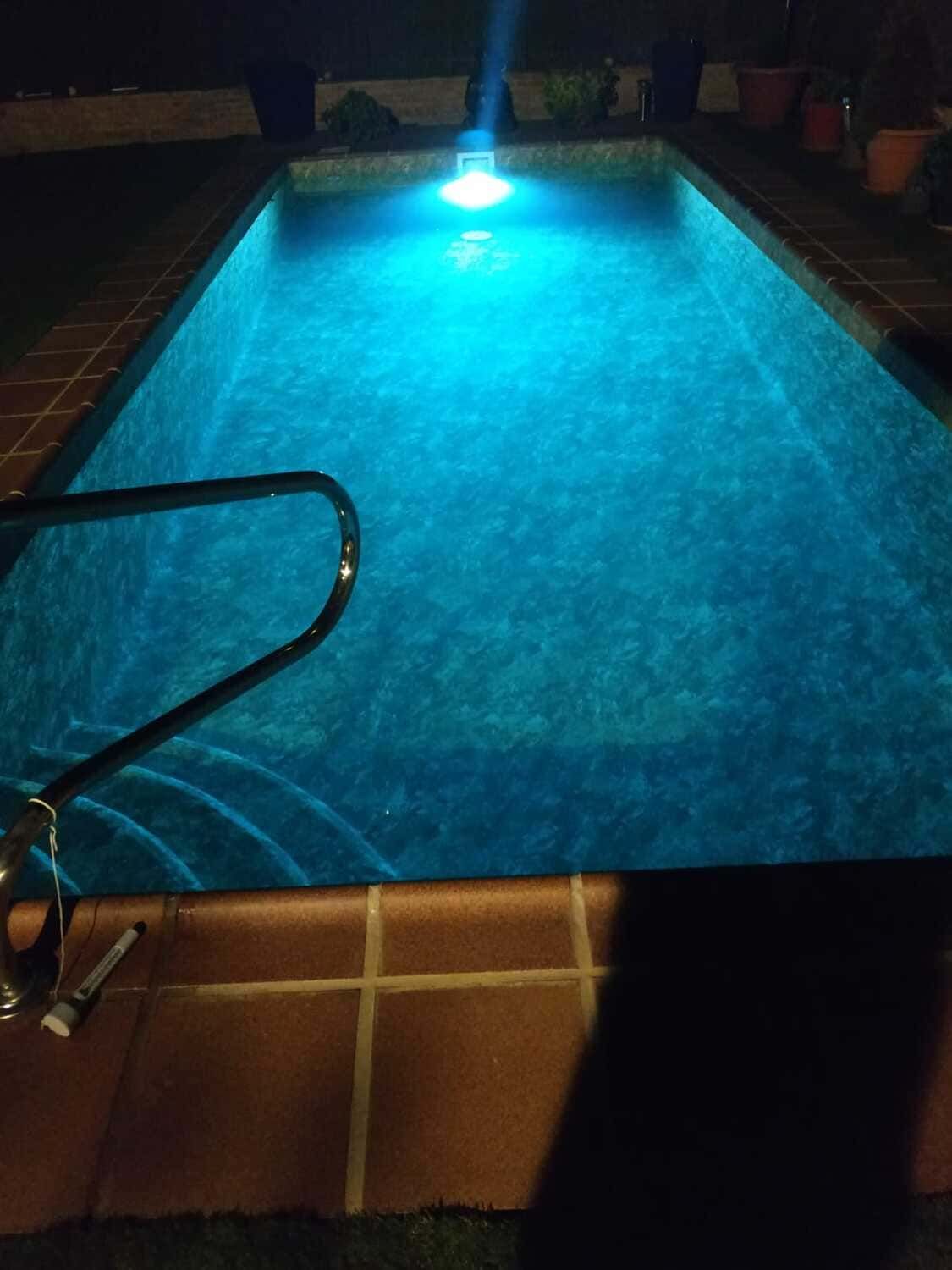 Vista nocturna de una piscina rectangular iluminada con luz azul. Se ve una barandilla metálica.