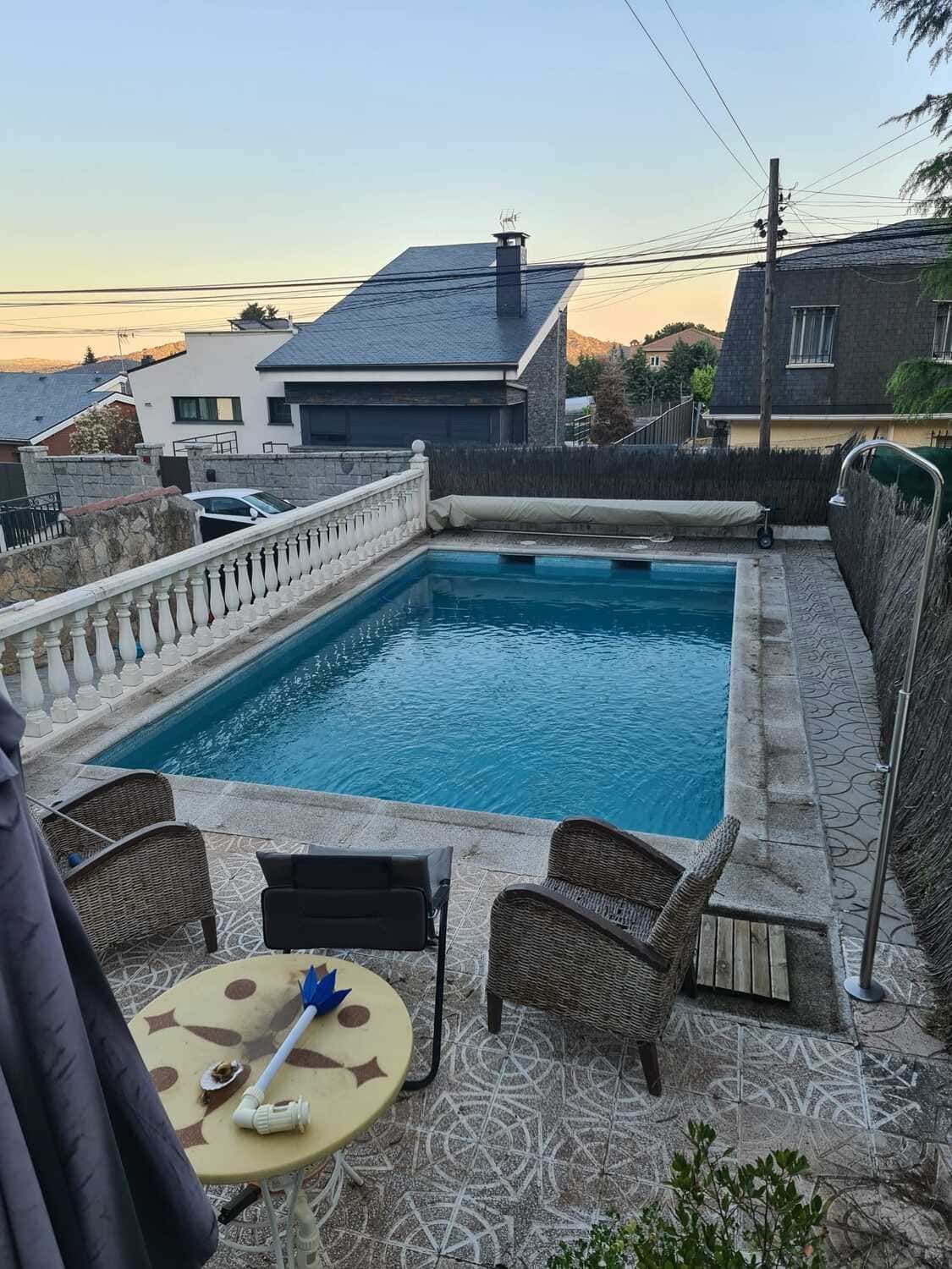 Piscina rodeada de patio de piedra, sillas y una pequeña mesa; casas y cielo al fondo.