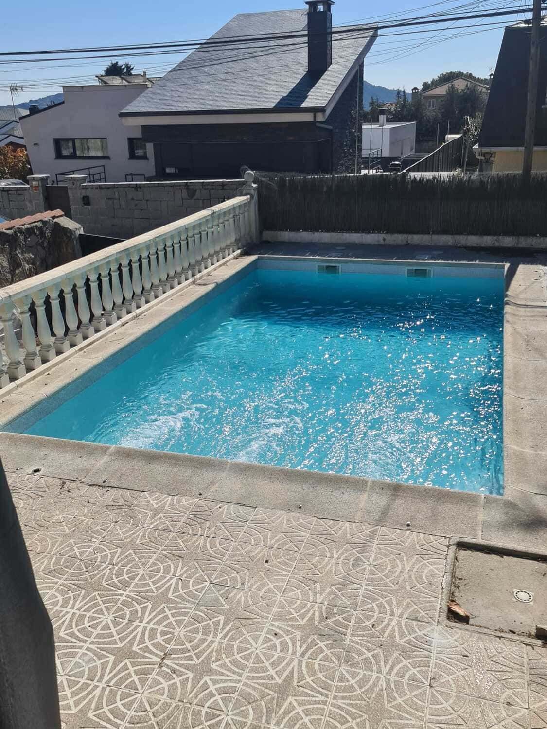 Piscina llena de agua azul, patio de hormigón, barandilla blanca y edificios al fondo.