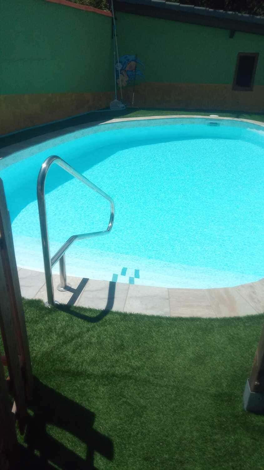 Piscina de agua azul cristalina con barandilla metálica, rodeada de césped artificial y pared verde.