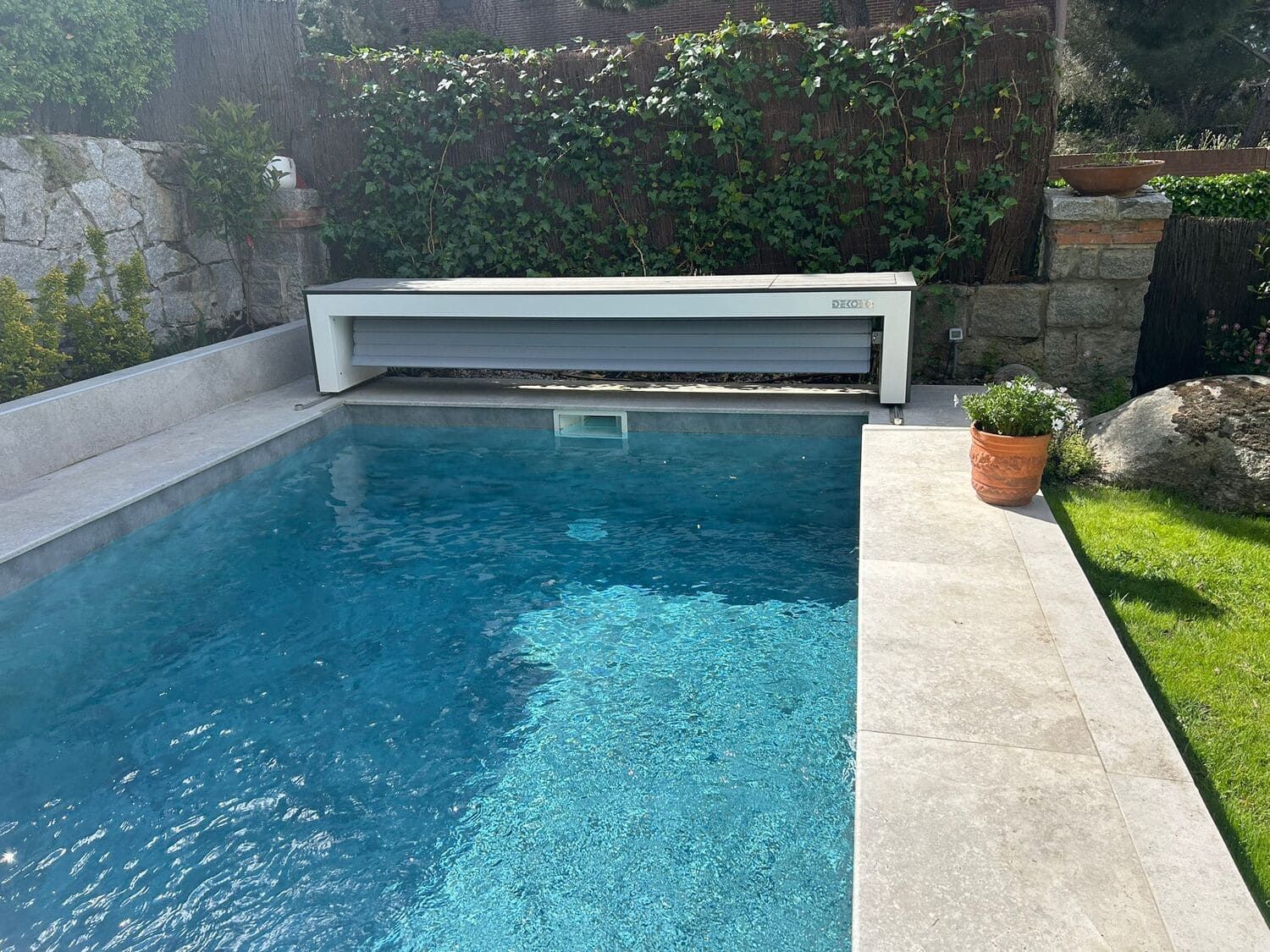 Piscina con cubierta blanca cerrada, rodeada de piedra y vegetación. En un entorno exterior soleado.
