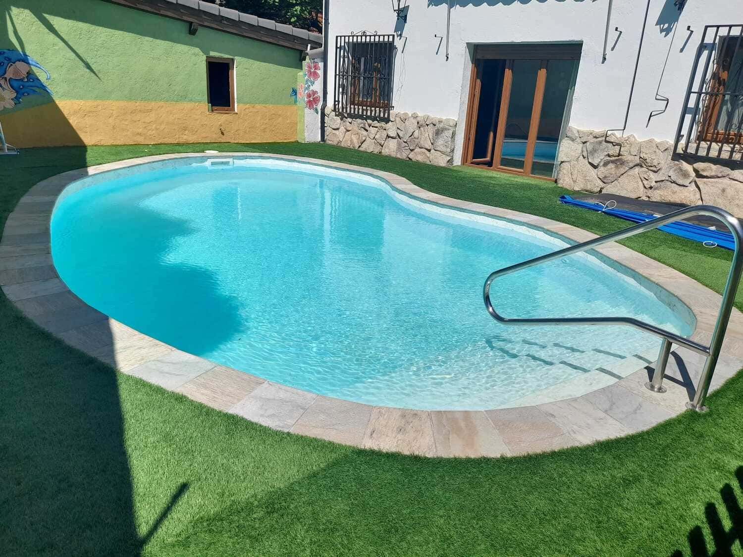 Piscina de forma ovalada, de agua azul, rodeada de césped artificial verde y borde de piedra.
