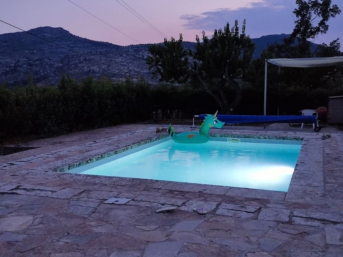 Piscina al anochecer con flotador de unicornio, rodeada de patio de piedra, árboles y fondo de montaña.