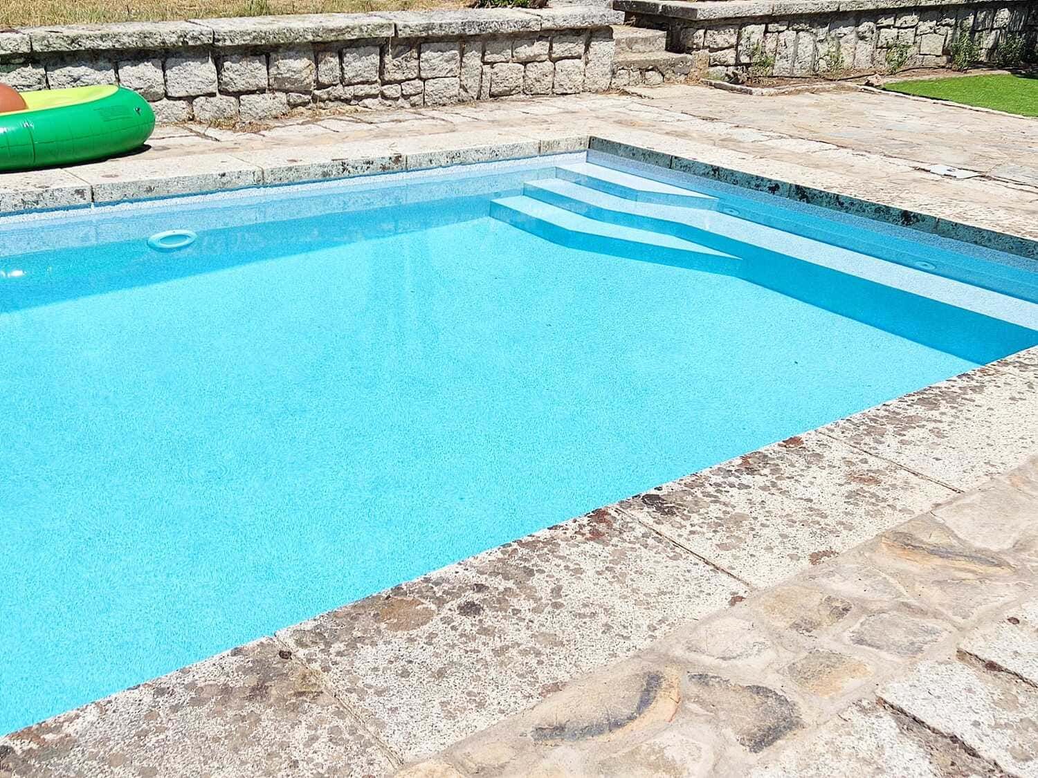 Piscina con agua azul y borde de piedra, en la que se ve un juguete inflable de color verde.