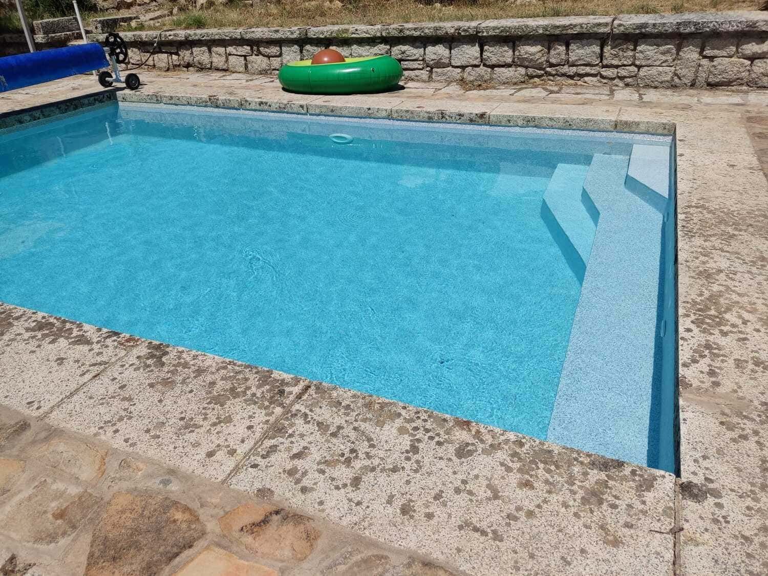 Piscina de agua azul con escaleras, rodeada de piedra. Cerca hay un inflable verde.