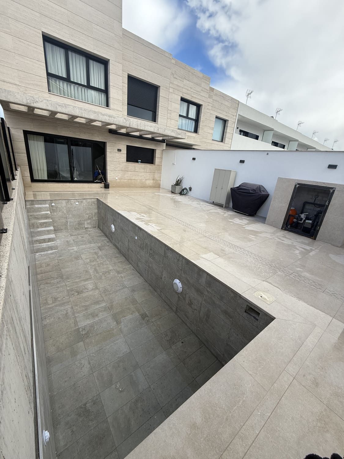 Piscina rectangular vacía en un patio. Casa moderna de color claro al fondo. Cielo nublado.