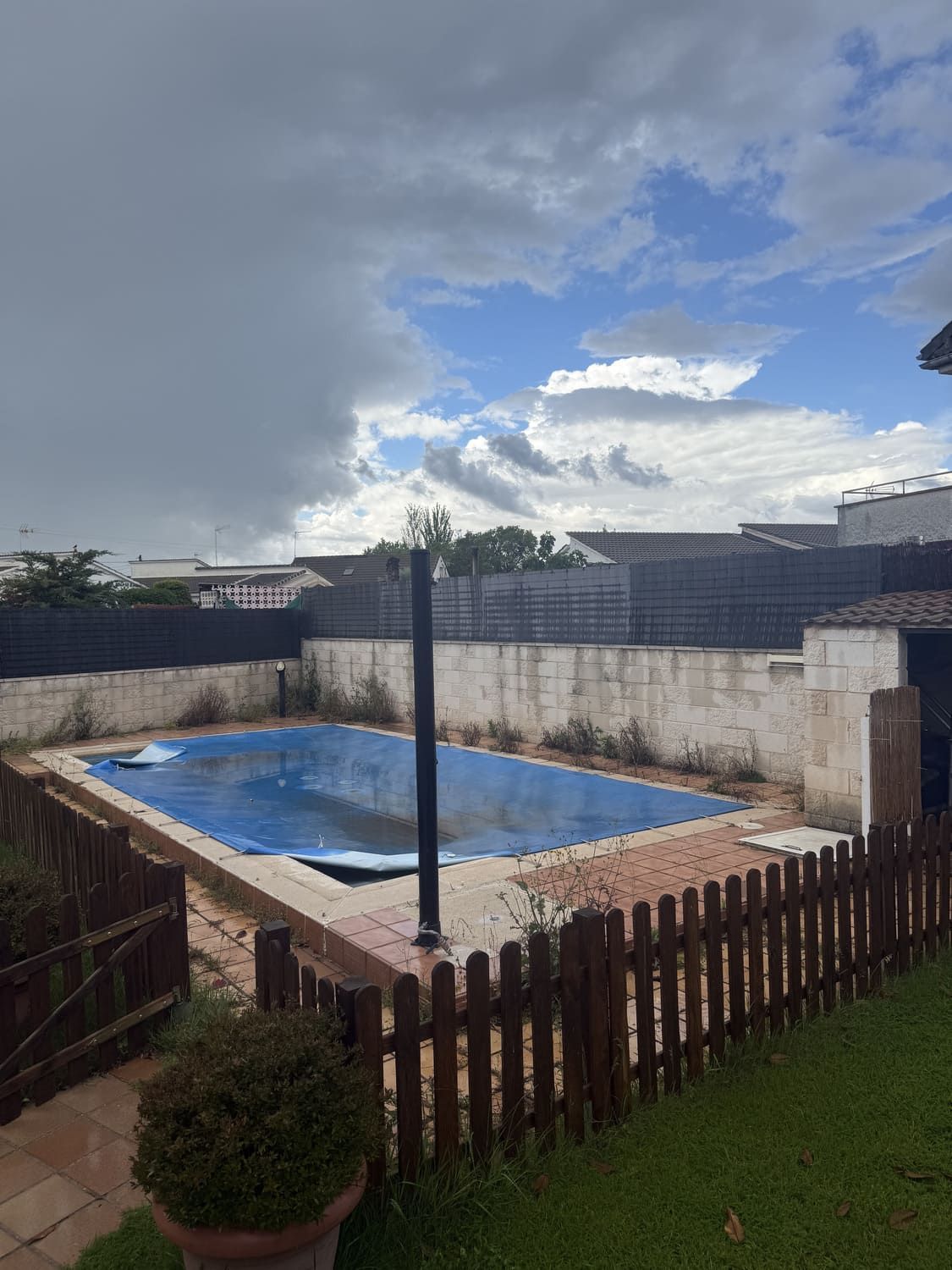 Patio trasero con piscina rectangular cubierta con lona azul, rodeada de una valla marrón y un muro beige. Cielo nublado.