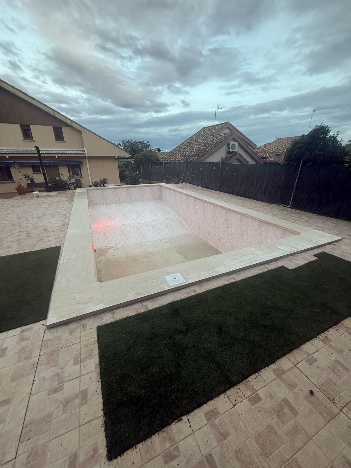 Piscina rectangular vacía con borde de piedra color canela, detalles de césped artificial y cielo nublado.